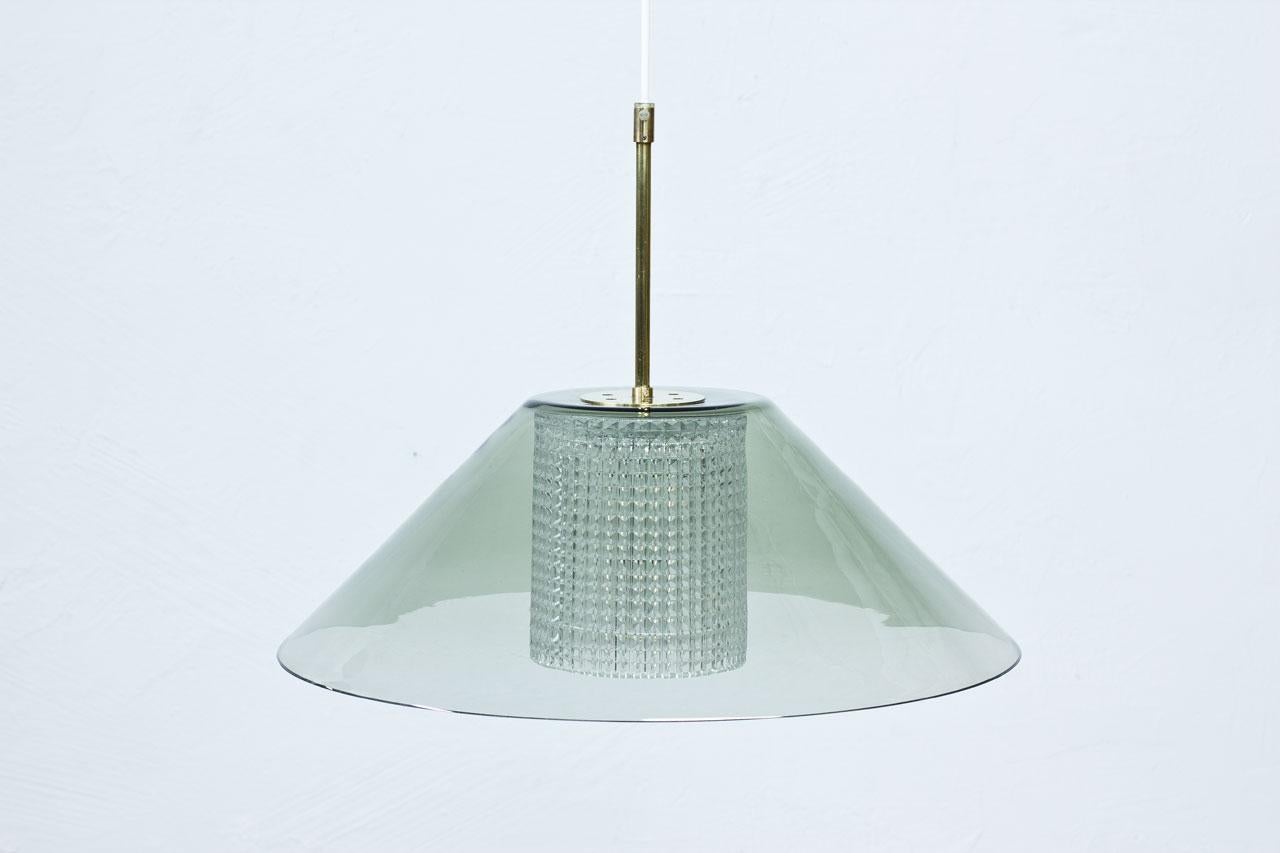 Pendant lamp designed by Carl Fagerlund for glass company Orrefors. Manufactured in Sweden during the 1960s. Outer cup in clear grey tinted glass with a cylindrical internal diffuser in clear pressed glass. Polished brass fittings. New electricity.