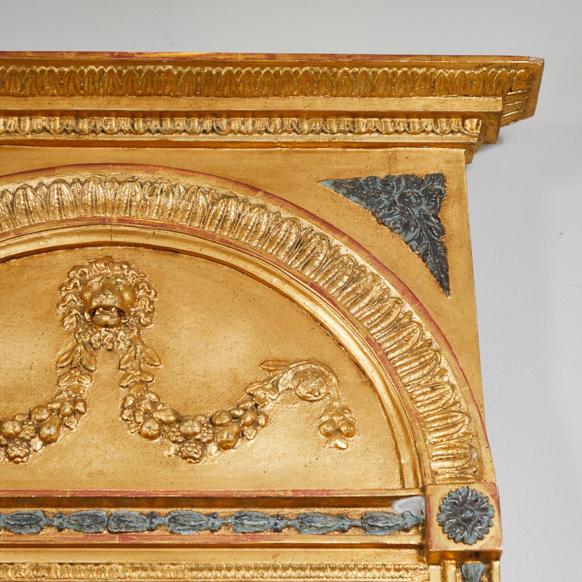 A Gustavian rectangular gilt mirror with beveled cornice top, carved arch frieze and anthemion accents. The neoclassical carving of a lion's head holding swags of fruit adds a regal touch. Painted blue architectural accents lend contrast and enhance