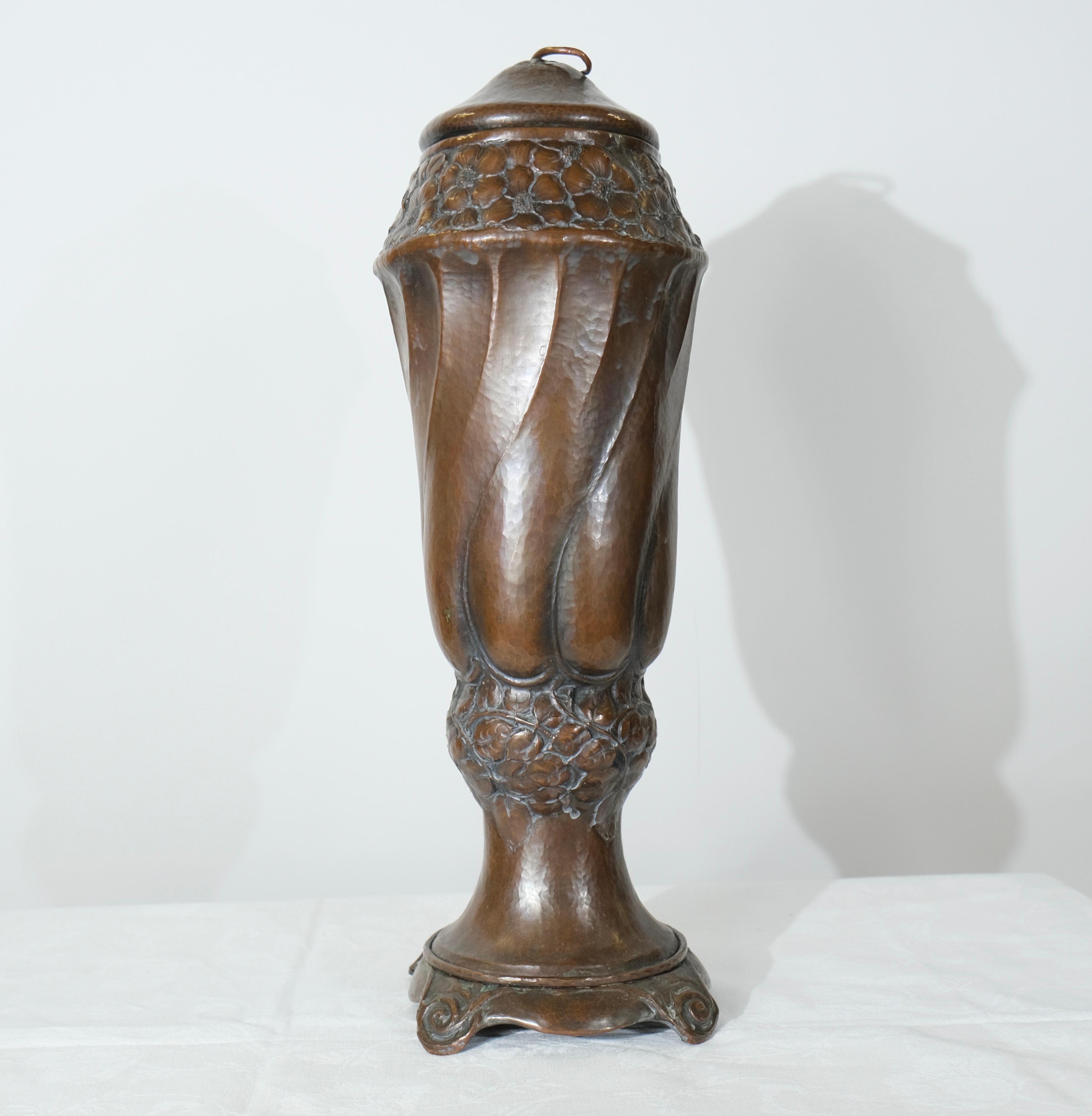 Questo vaso in rame con coperchio incarna lo stile Art Nouveau, caratterizzato da linee organiche e fluide e da dettagli intricati. Il vaso presenta una forma allungata e contorta con motivi vorticosi simili a petali che creano un senso dinamico di