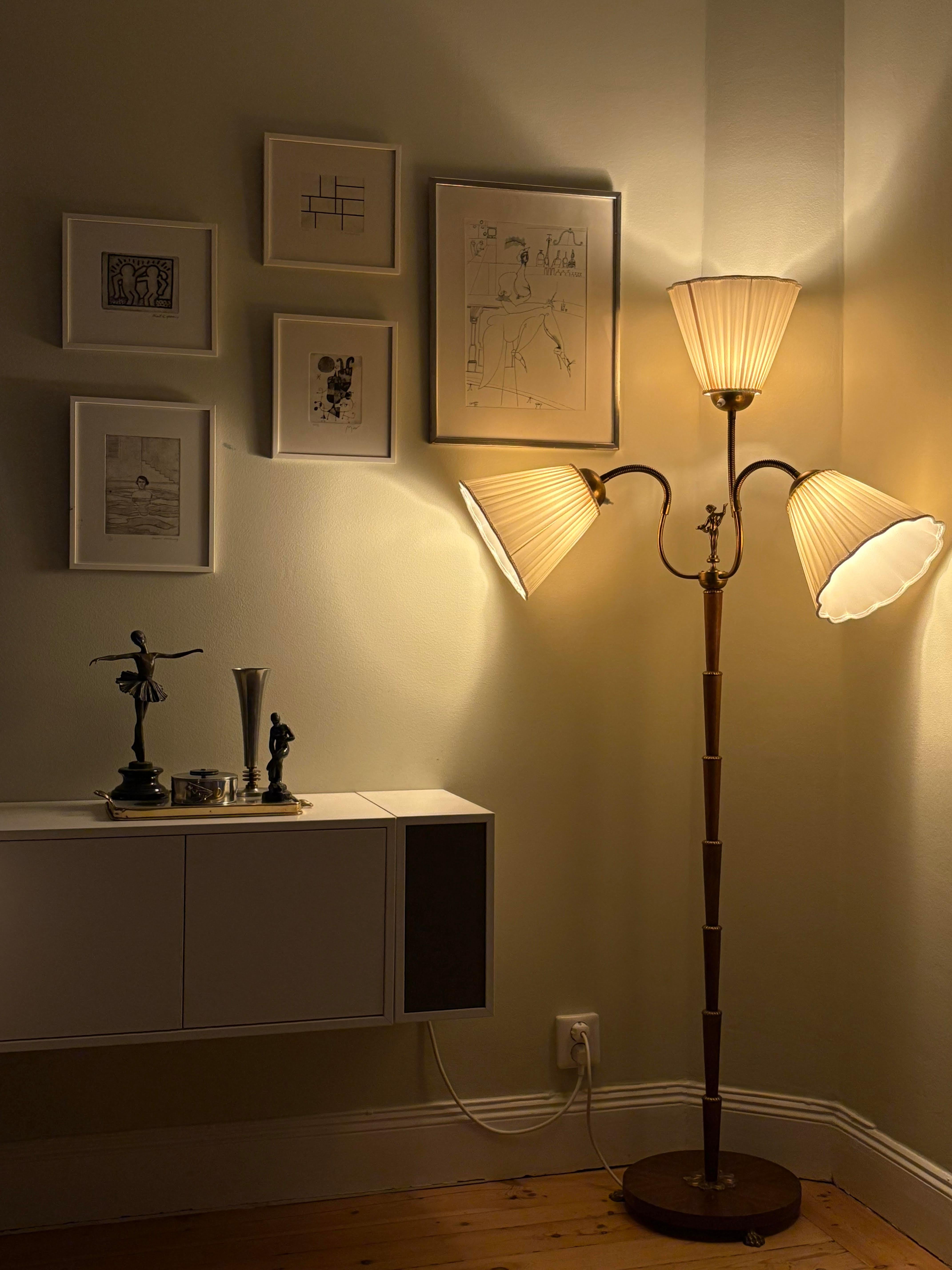 A charming mid-century floor lamp from Sweden, dating to the 1930s. This elegant piece features a finely segmented wooden stem rising from a circular base, with three gracefully curved brass arms that support pleated silk lampshades. Each shade