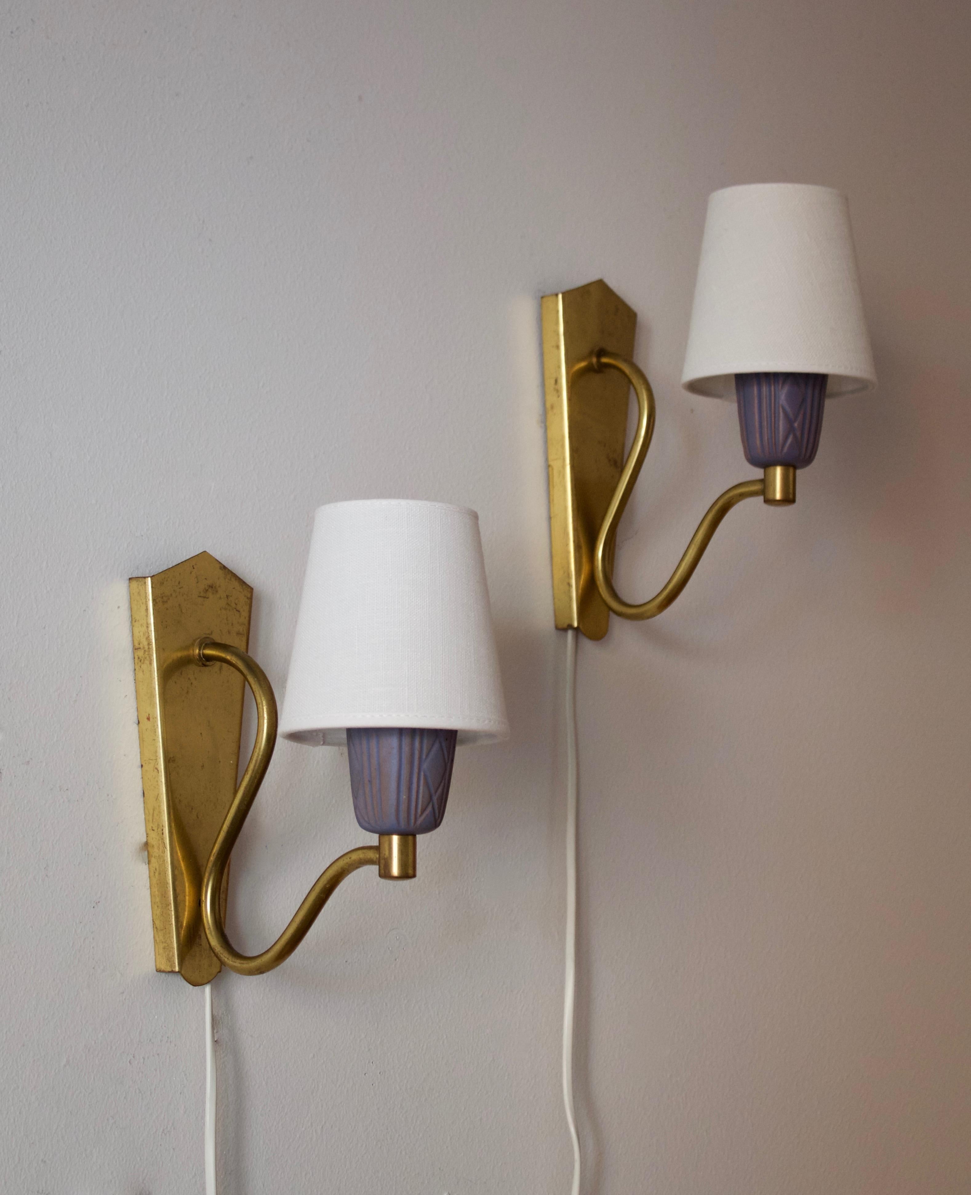 A pair of small wall lights. Designed and produced in Sweden, 1950s. Features brass, blue glazed ceramic sockets with incised decoration. Brand new linen lampshades.