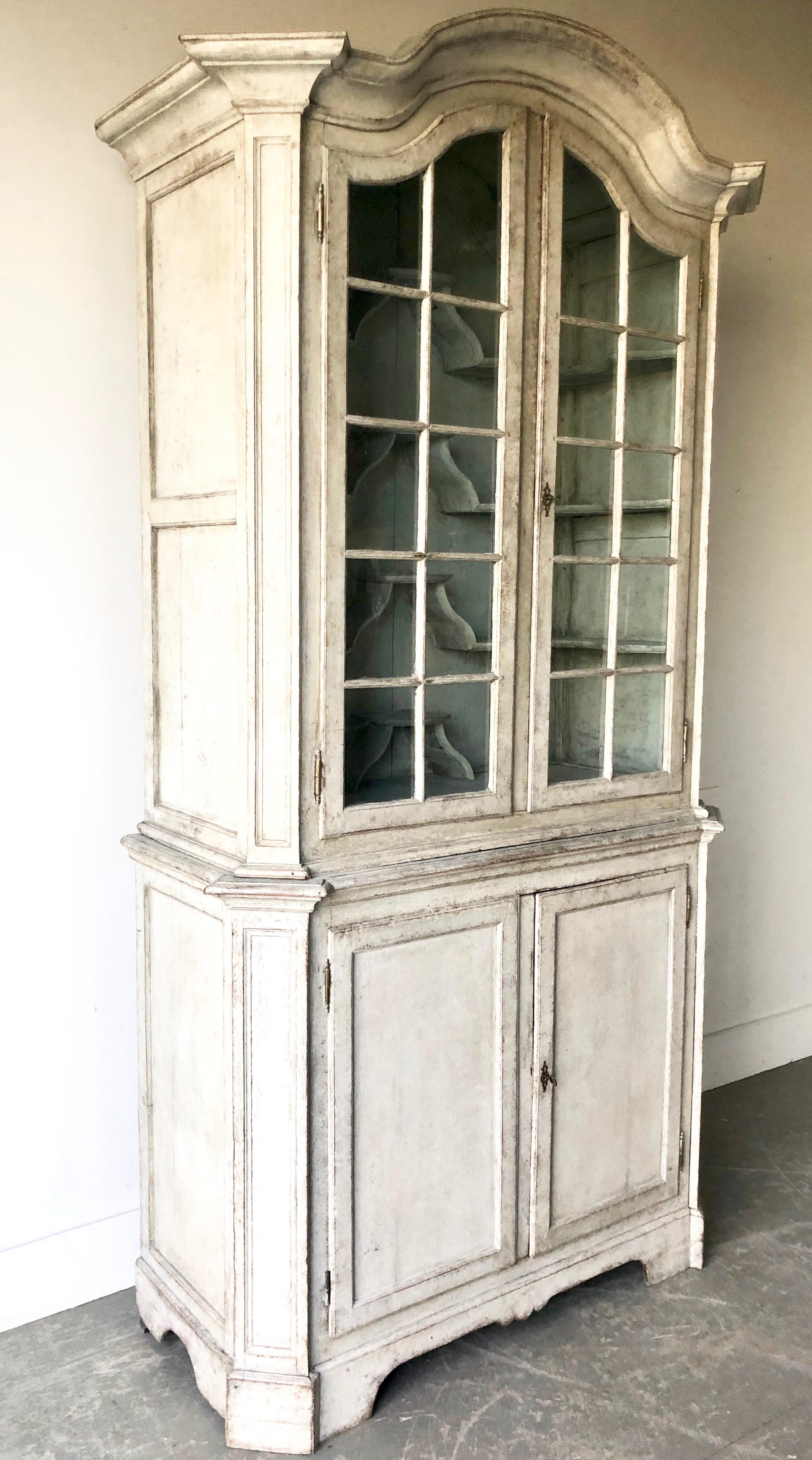 Swedish early 18th century period Rococo Vitrine cabinet in two parts with arched pediment over original glazed panel doors with multiple unusual bracket display shelves, supported the sturdy two-door base with bracket feet. Painted in grayish