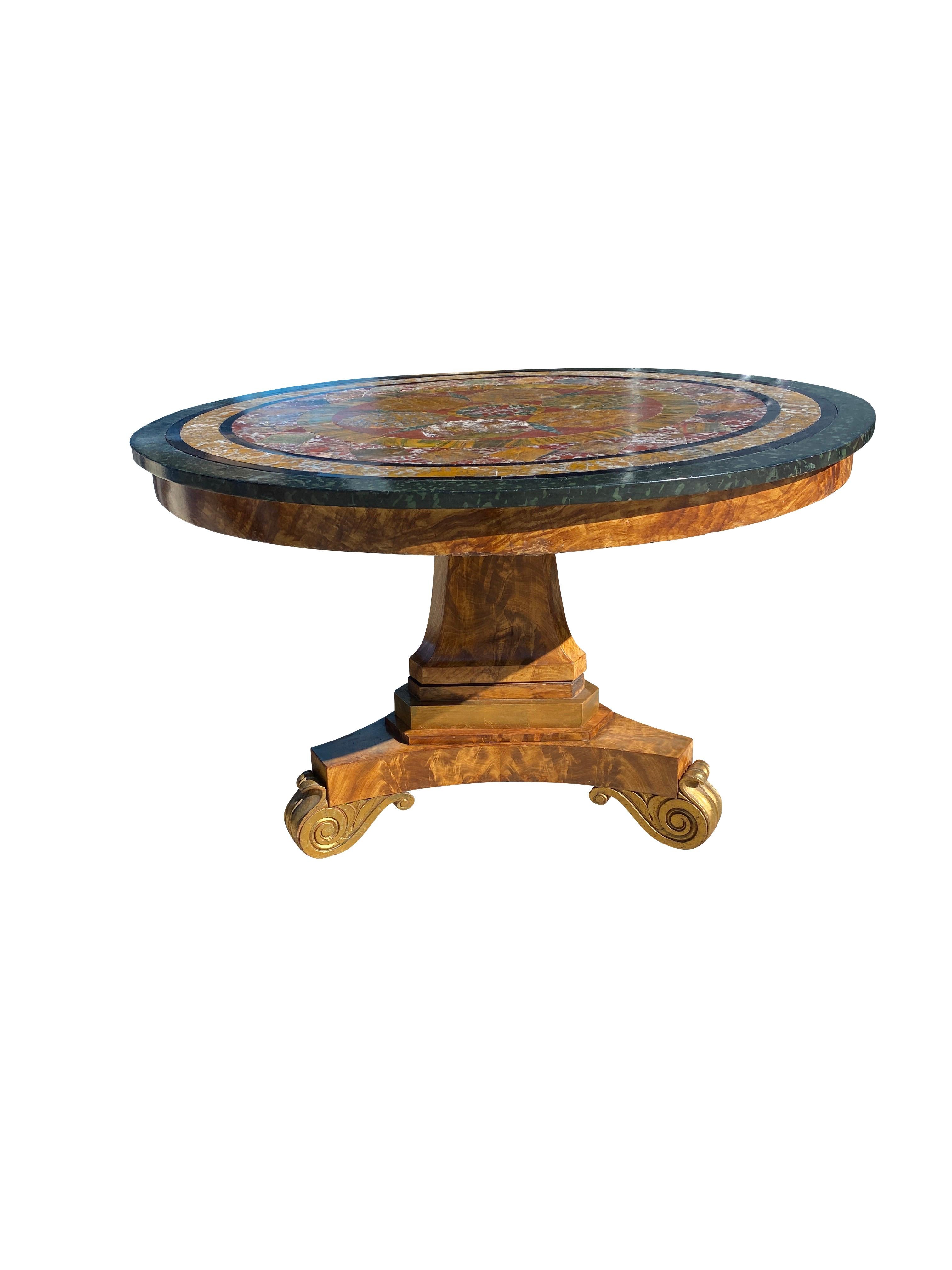 Circular top with wood edge surrounding a geometric mosaic of hard stones raised on a triangular tapered column and tripartite base with gilded scroll feet. From the estate of William Hodgins a noted Boston interior designer. The piece is featured