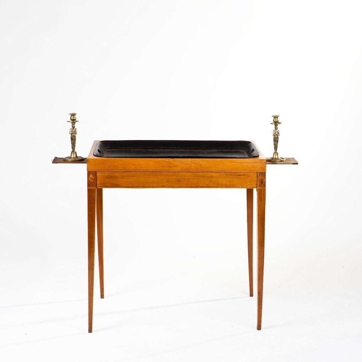 Elegante mesa bandeja Imperio Sueco con candelabros extraíbles, fabricada en abedul con incrustaciones de rombos en arce y cordones de madera clara satinada a lo largo de los faldones y las esquinas.
Las patas están finamente ahusadas, lo que