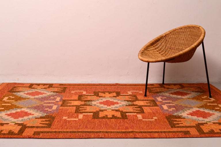 A flat-weave rölakan rug / carpet designed by Ingegerd Silow, Sweden. Hand woven with a geometric pattern where the colors are different shades of rust red, and orange, purple, brown, and some beige, blue, yellow and green. This carpet is in a very