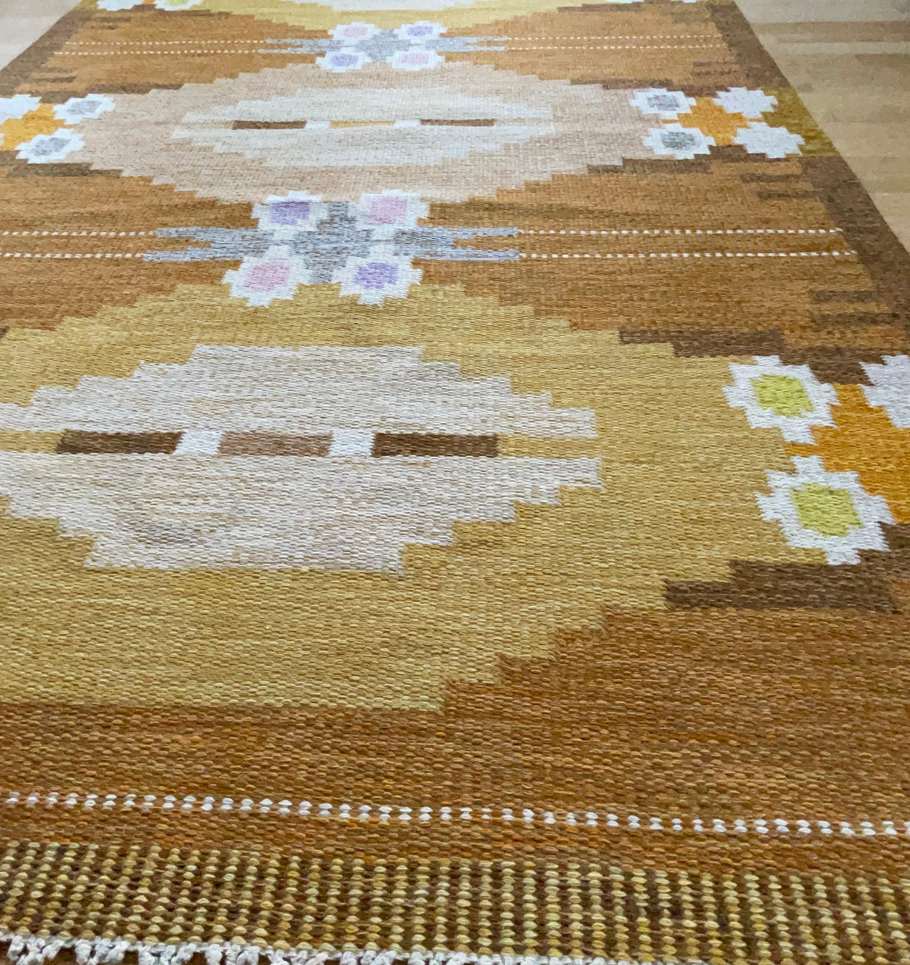 A flat-weave rölakan rug / carpet designed by Ingegerd Silow, Sweden. Hand woven with a geometric pattern where the colors are different shades of burnt orange, mustard yellow, and some blue, beige, purple and pink. This carpet is in a very good