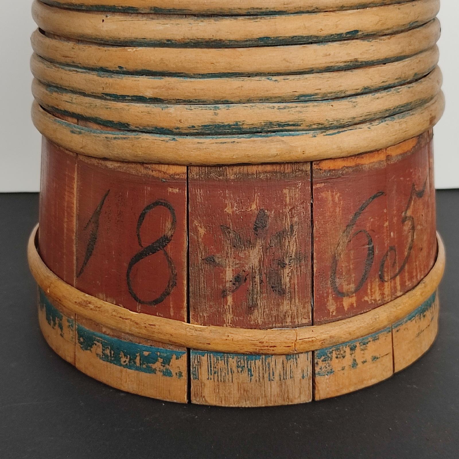 Norwegischer Holzkrug aus dem 19. Jahrhundert, bemalt mit Initialen und datiert 1865.
Holzkrug mit Deckel, hergestellt aus Dauben, die mit einer Banderole aus gespaltenen Ästen zusammengehalten werden. Die Deckel sind mit Scharnieren versehen.