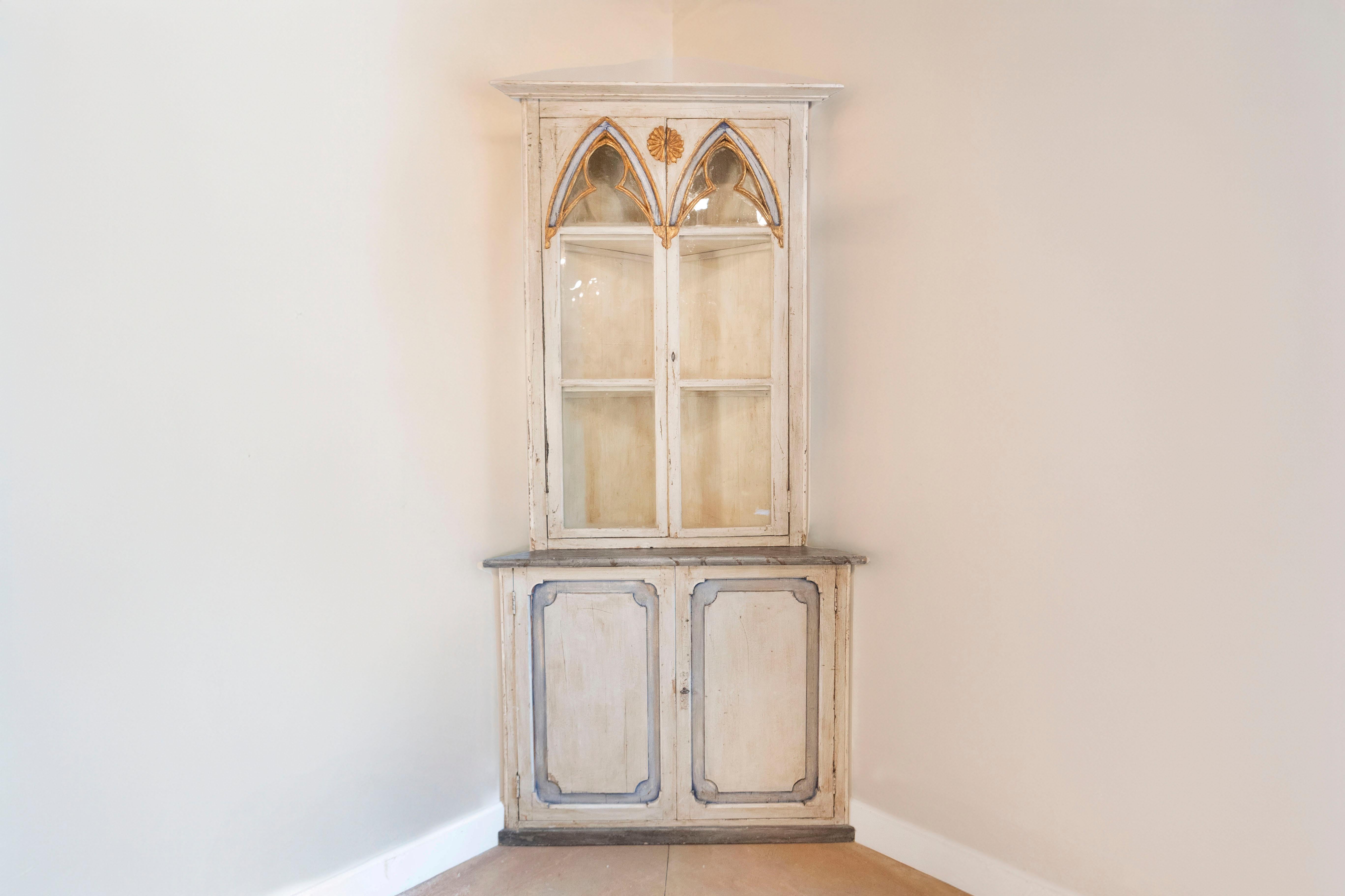 A Swedish Gothic Revival painted wood and glass doors corner cabinet from the mid-19th century. This exquisite Swedish corner cabinet features a molded cornice overhanging two Gothic inspired doors with glass panels. A giltwood rosette, carved onto
