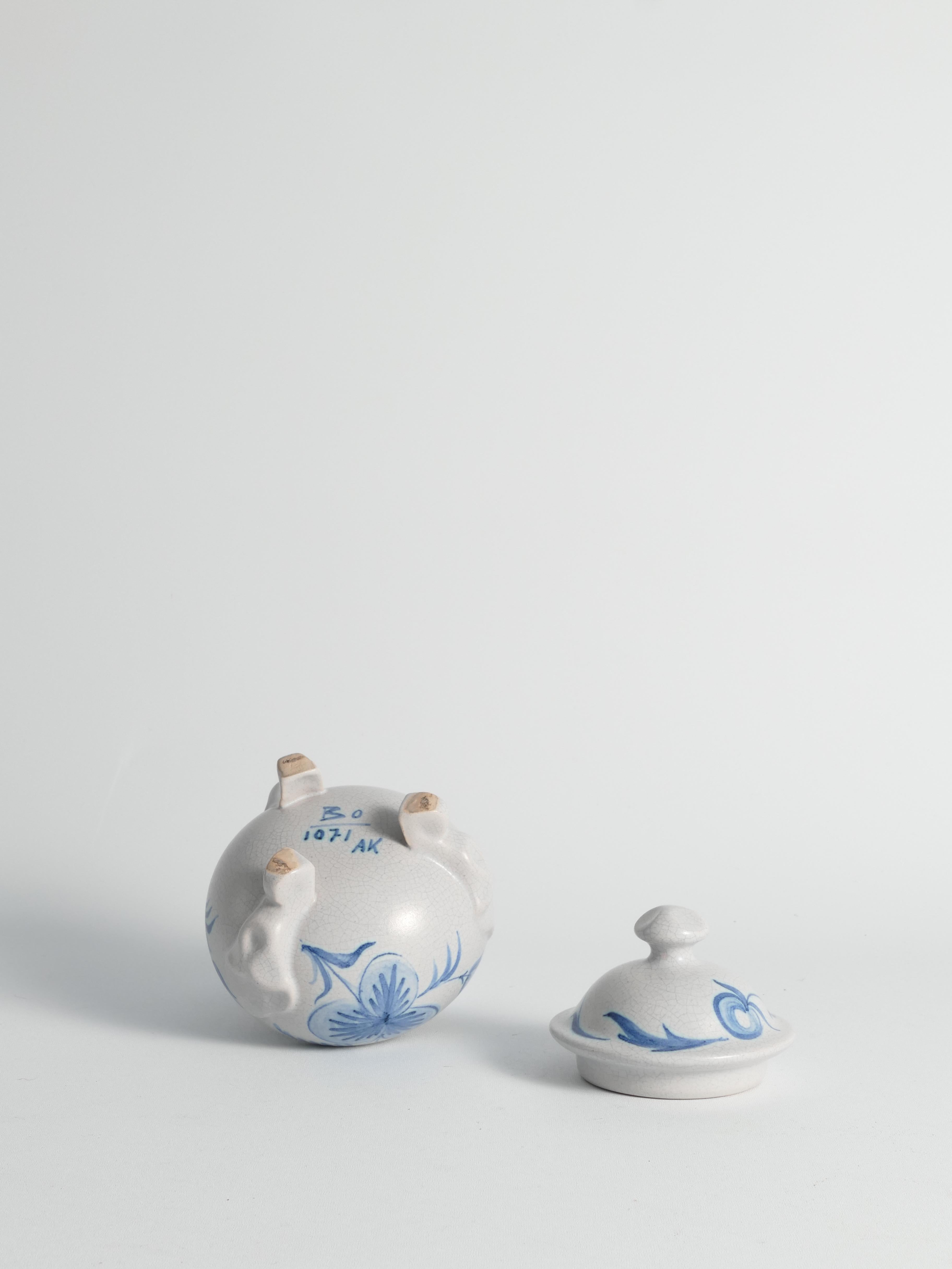 Questa deliziosa bomboniera con coperchio è stata disegnata da Allan Ebeling per Bo Fajans,  Si erge con grazia su tre piedi curvi, emanando fascino con la sua forma globosa. La sua tonalità grigio chiaro, ornata da un delicato decoro floreale blu