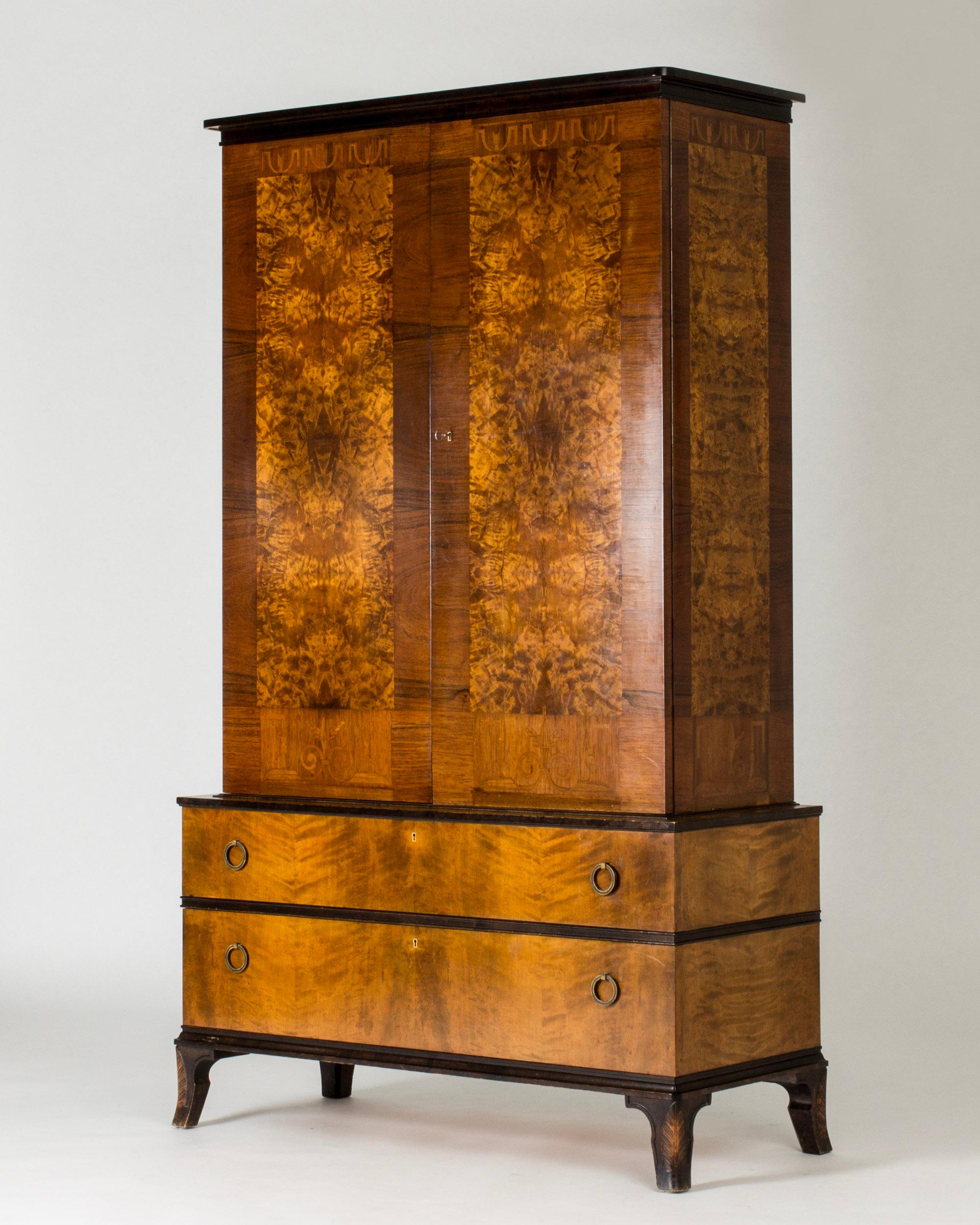 Elegant mahogany cabinet by Erik Chambert, with drawers in the wider bottom part. Dramatic root veneer on the cabinet doors, decor of intarsia. Round brass handles. Legs with carved pattern of feathers.