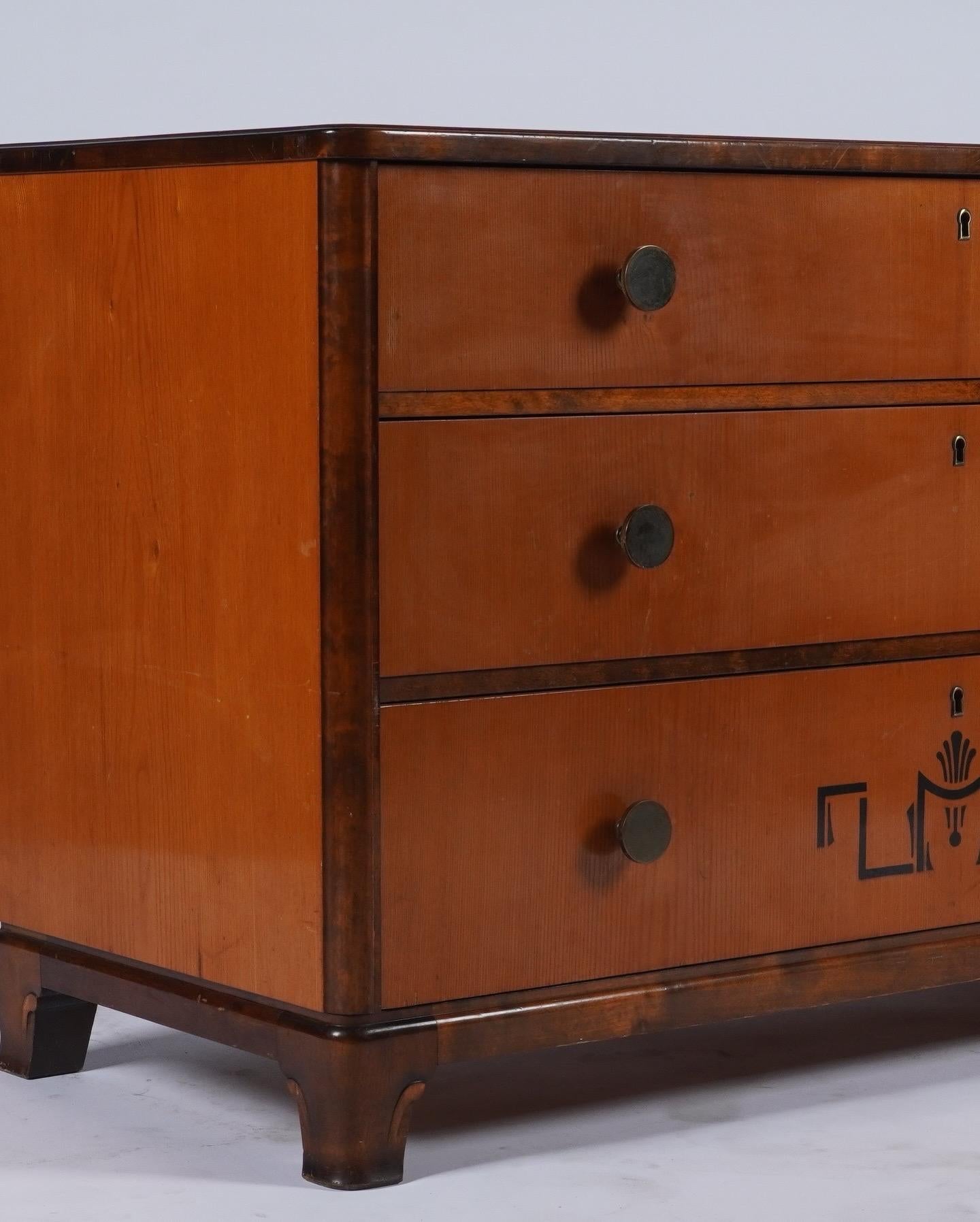 A Swedish grace or Danish art deco commode in solid elm trimmed with flamed birch. Intarsia decorates the lowest of the three drawers. Patinated brass pulls. Drawers are deep and slide smoothly. The cabinet retains its original finish with