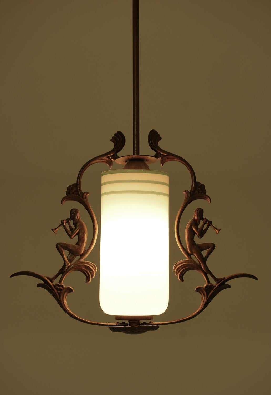 A Swedish Grace pendant lamp from the 1930s with a bronze-patinated frame and an opaline glass shade. The design features two decorative figures playing trumpets, adding a subtle artistic detail. The cylindrical glass shade has green stripe accents