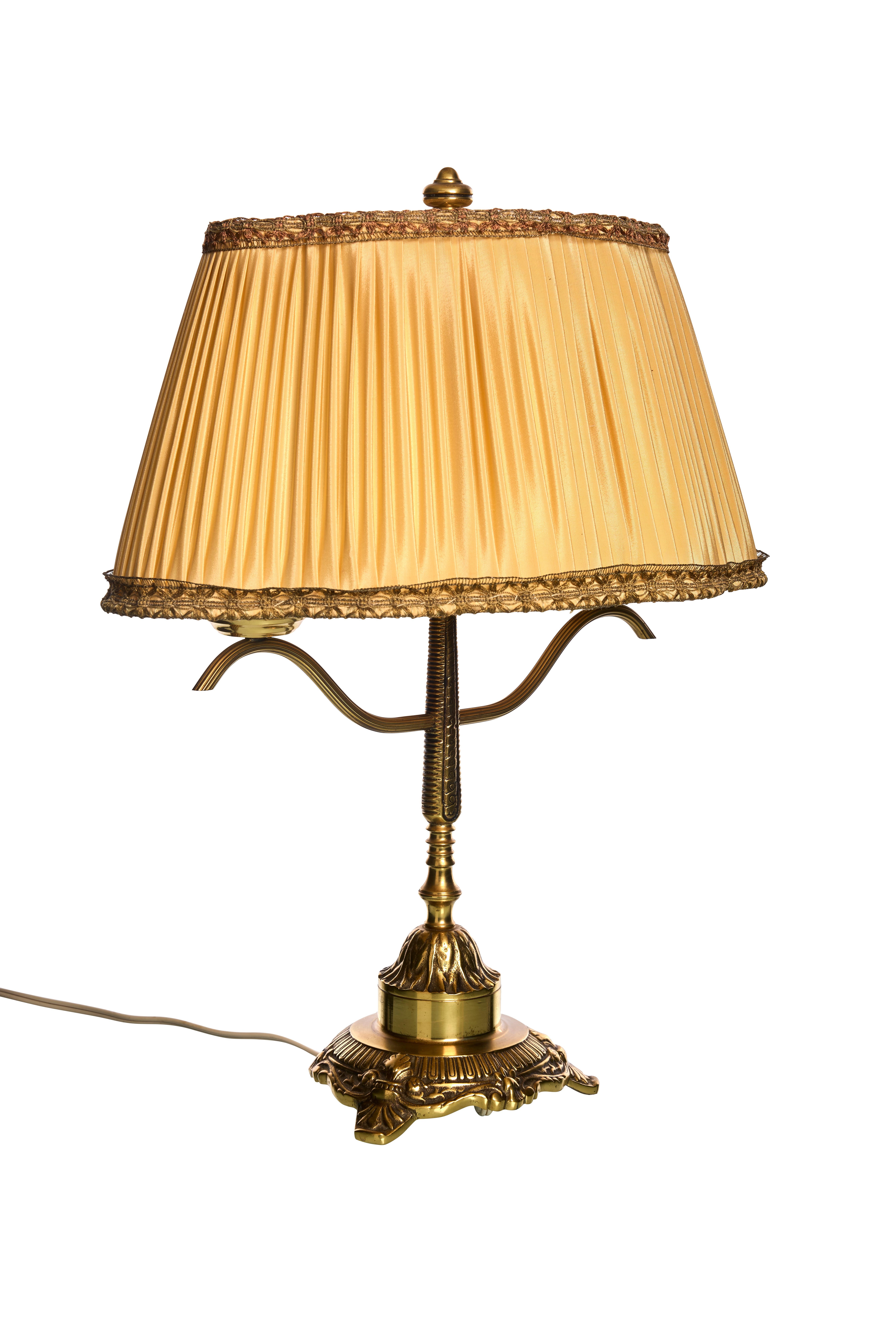 This table lamp features a classic design with a warm gold tone. The base is ornately detailed with intricate patterns, lending an antique feel. The lamp is topped with a pleated golden fabric lampshade, edged with decorative trim, enhancing its