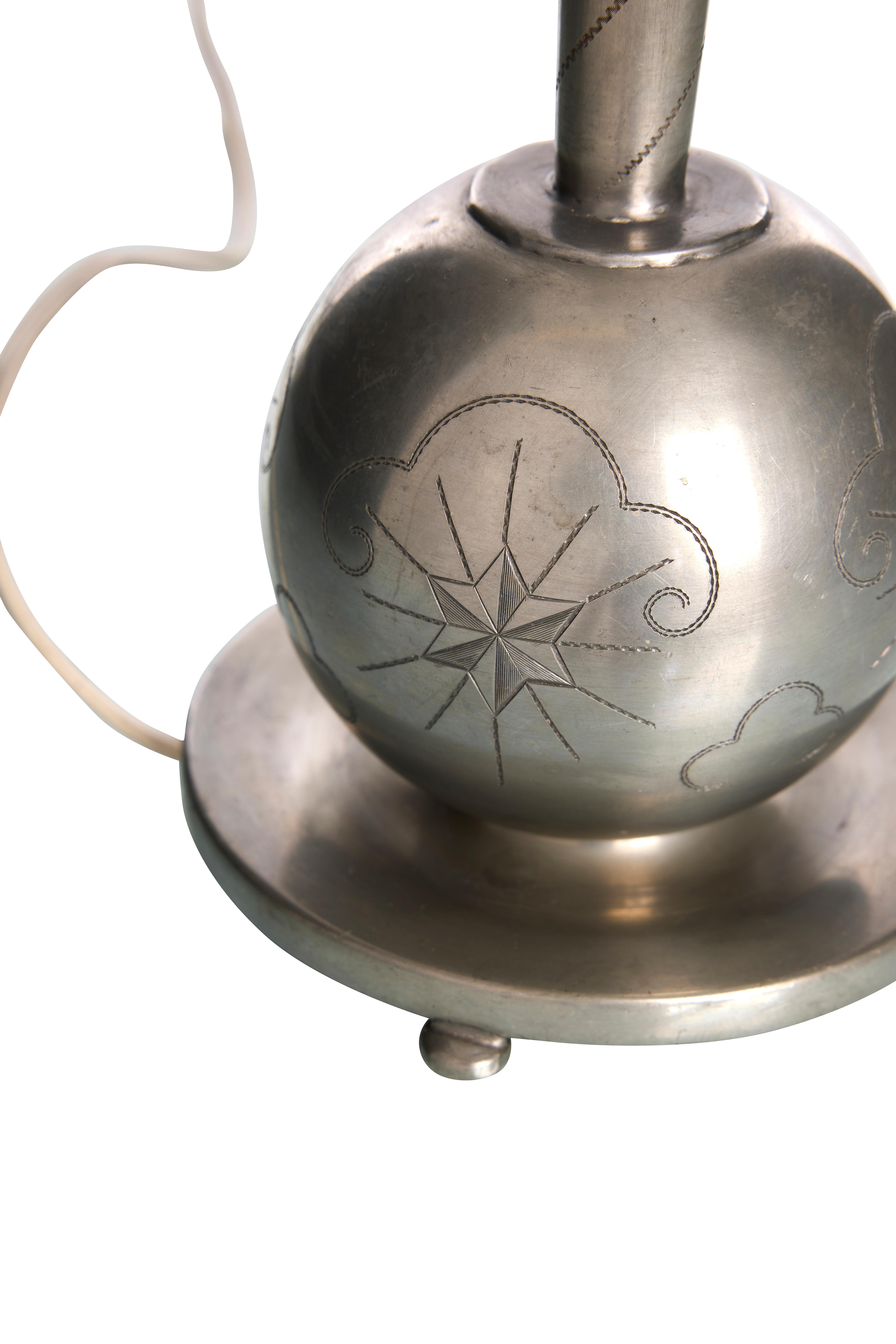 This is a vintage table lamp from the 1930s designed by Celestine Andersson for Cela. It features a sleek, metallic finish with intricate engravings on its spherical base. 

The design showcases elegant, swirling patterns and a prominent star motif,