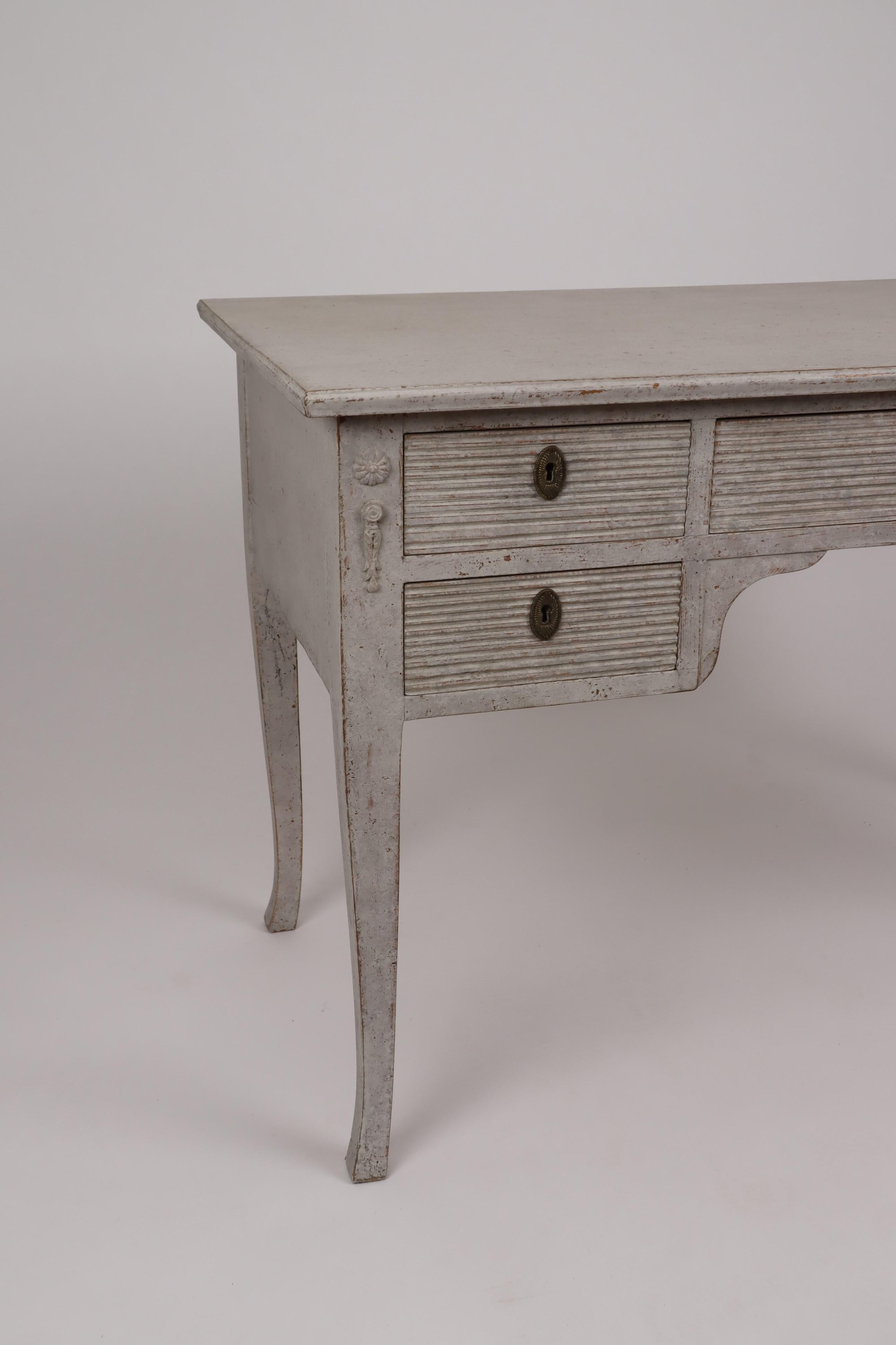 A Swedish five drawer desk from the 20th century with gray beige painted finish, carved reeded accents and slightly curving legs. Discover the understated charm of this Swedish 20th-century desk, presenting a harmonious blend of utility and Nordic