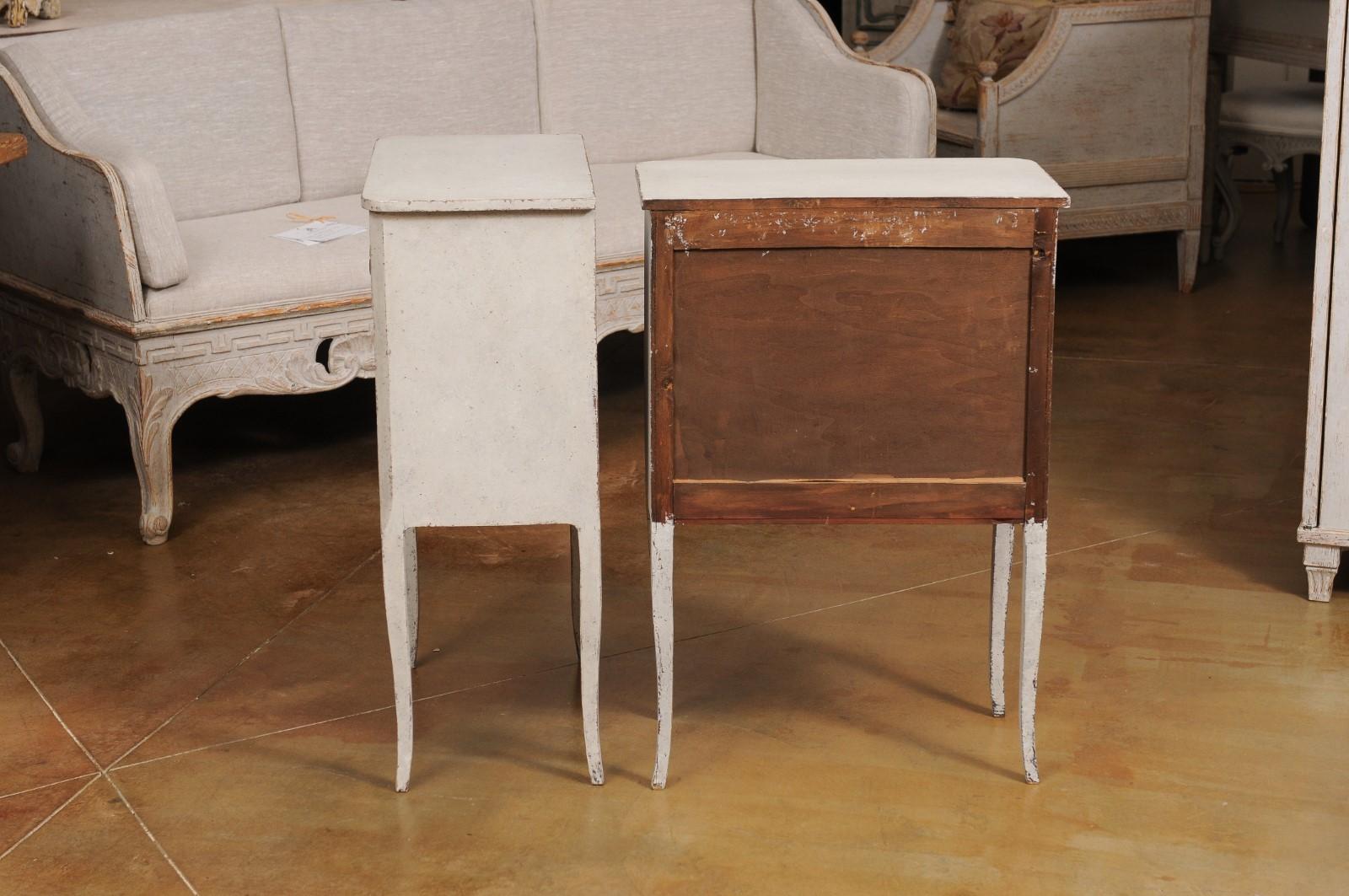 A pair of Swedish bedside tables from the 20th century with light gray painted finish, three drawers, ornate hardware, curving legs and carved aprons. Embodying the quintessence of Scandinavian elegance, this duo of Swedish 20th-century bedside