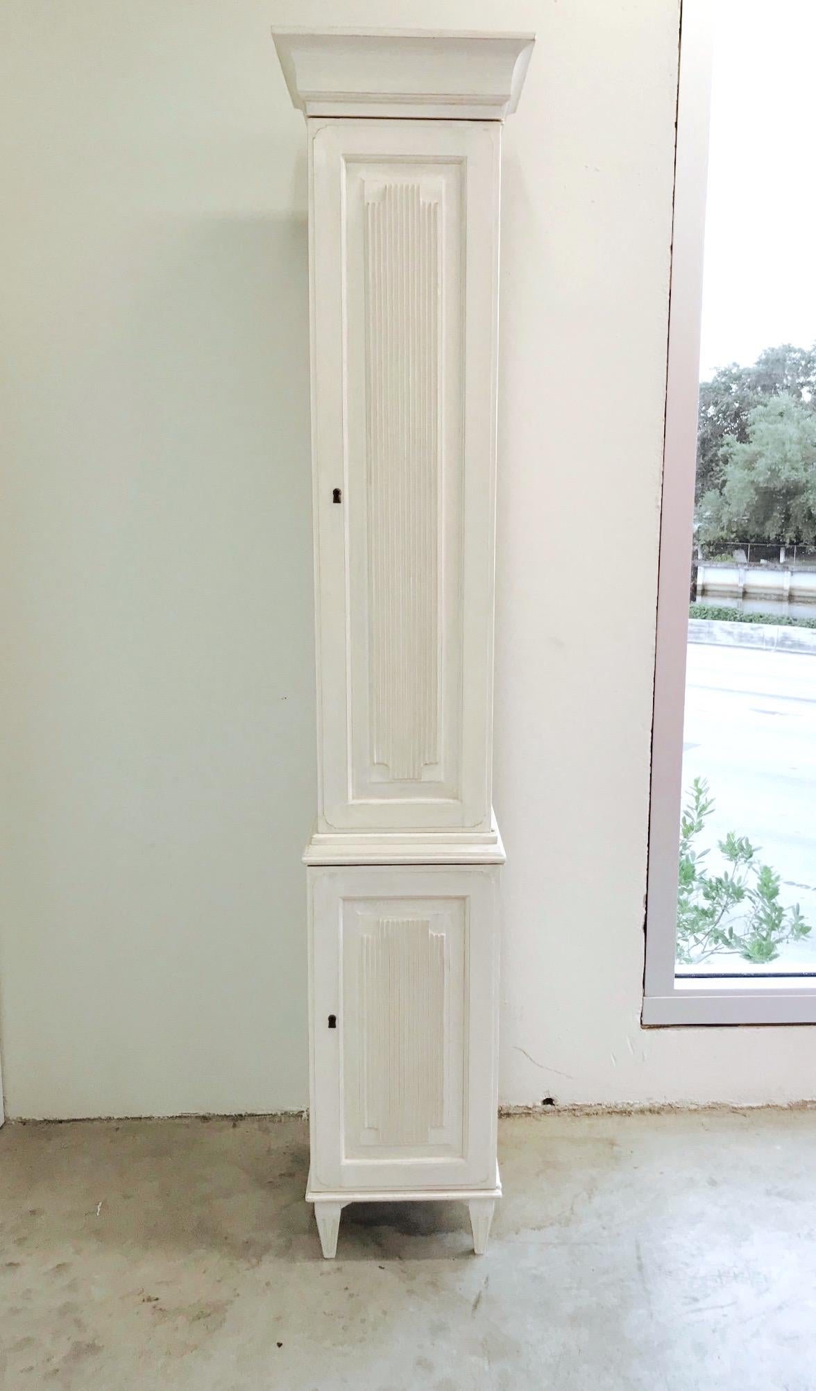 Exquisite Swedish carved wood tall and narrow cupboard with Gustavian style hand painted finish. The cabinet features incised wood carvings throughout. Features molded doors with fluted designs and neoclassical pediment top. Top cabinet is fitted