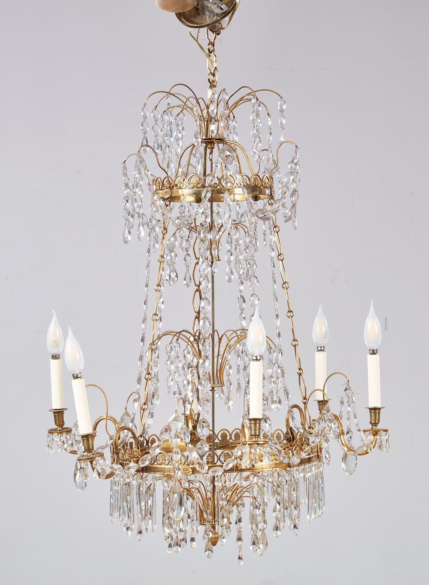 An elegant Gustavian crystal and gilt brass Swedish chandelier with two fountain-like tiers of faceted crystals over a central drop to an elaborate brass ring decorated with Egyptian heads and patera from which extend six curved arms repeating the