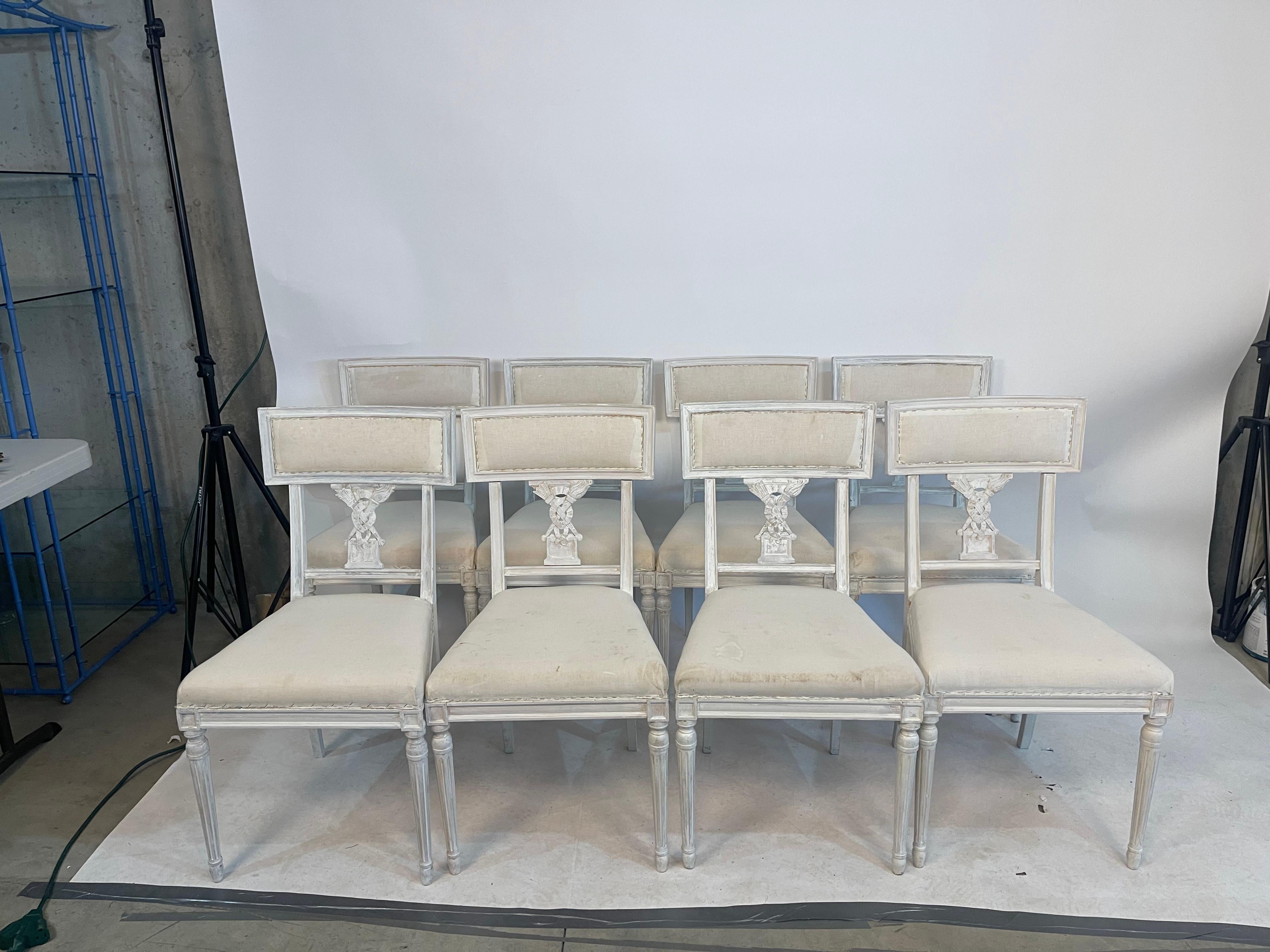 Set of 8 petite Northern European dining chairs in washed white wood paint with neoclassical decoration on the back splat. The decoration includes crossed flaming torches on a classical plinth. The all-over effect is a stylish classical chair