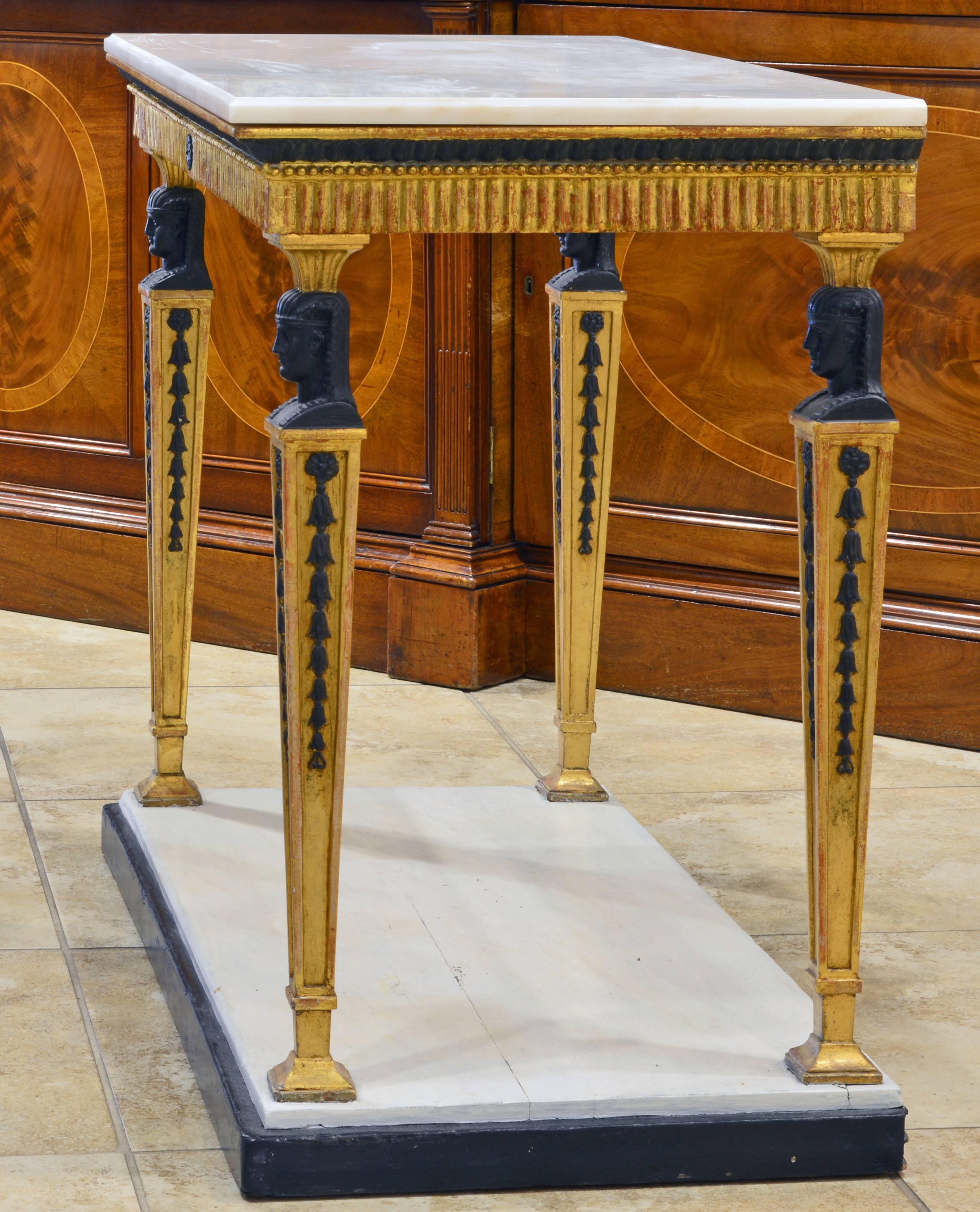 This refined Gustavian console table dating to the early 1800s features a white marble top with molded edge above a carved gilt-wood frieze finished on three sides and supported by four square tapering legs surmounted by ebonized pharaonic busts,