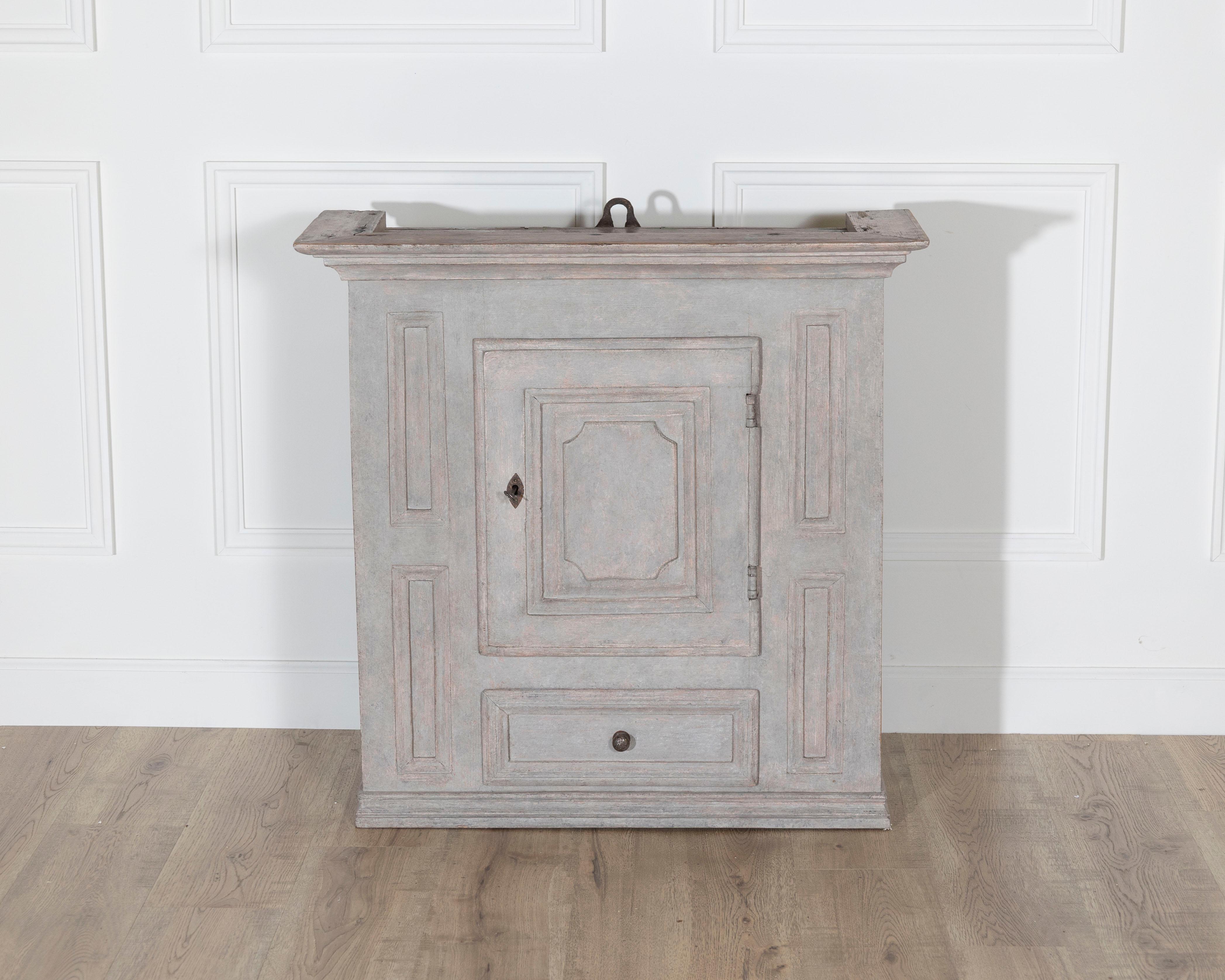 This charming Gustavian hanging kitchen cabinet, crafted in pine and finished in a soft grey patina, dates to the early 19th century. Its architectural structure features a single paneled door with molded trim and a lower drawer, both accented by