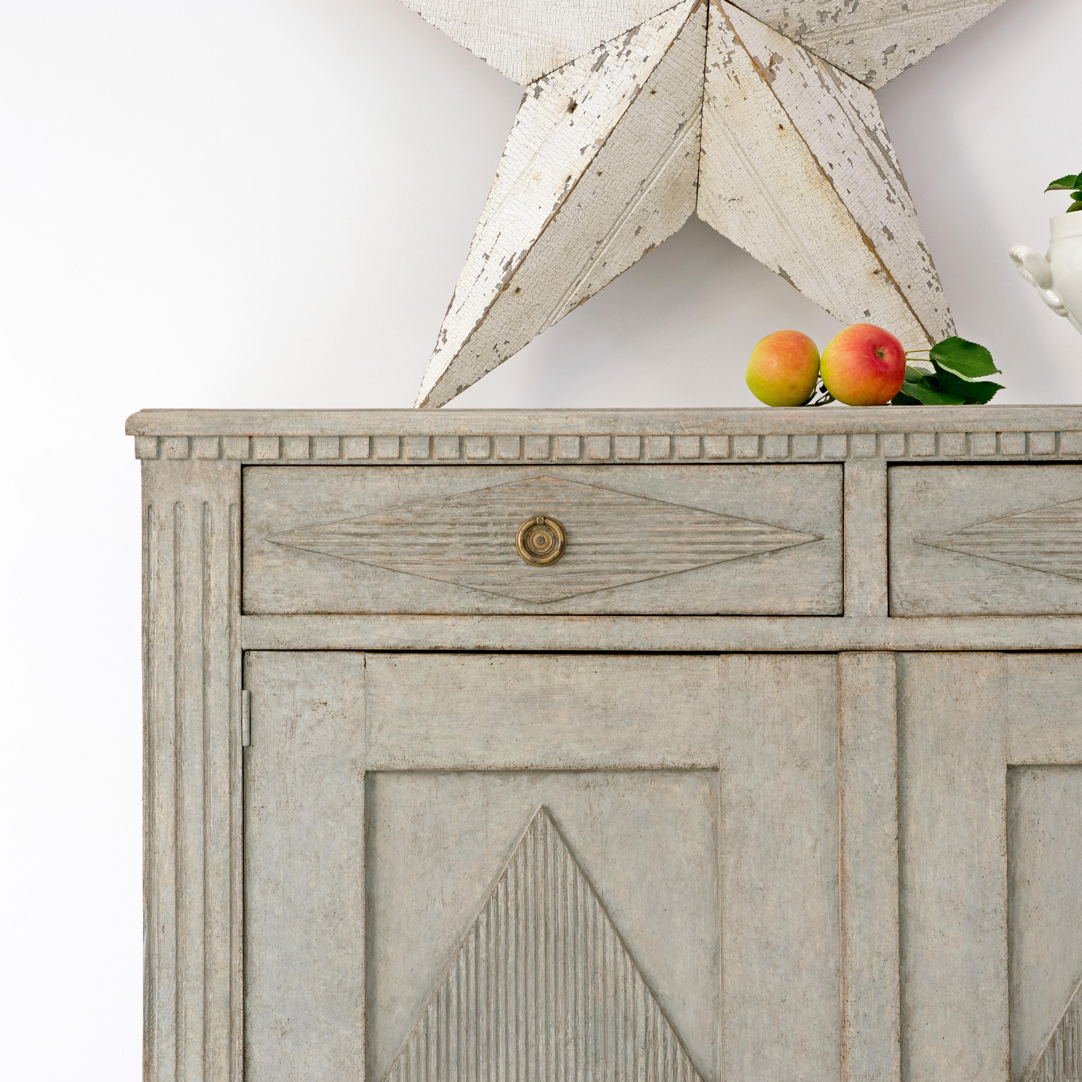 A Swedish Gustavian style painted wood sideboard circa 1840 with carved dentil molding, two drawers over two doors, carved and reeded diamond motifs and tapered feet. Embodying the refined elegance of the Gustavian style, this circa 1840 Swedish
