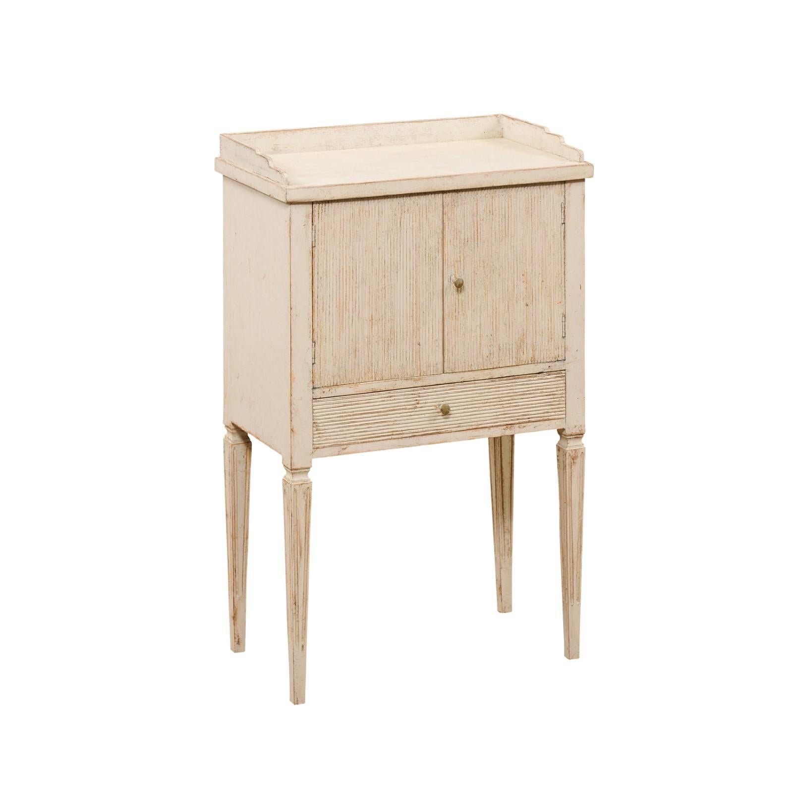 A Swedish Gustavian style painted wood nightstand from the mid 19th century with reeded doors, low drawer, carved gallery and tapered legs. Created in Sweden during the 1850s, this painted bedside table features a rectangular top with three-quarter