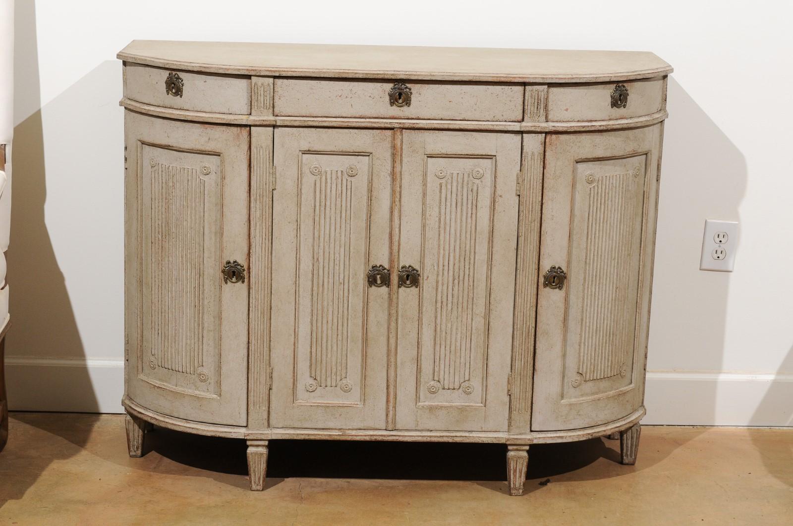 A Swedish Gustavian style painted demilune sideboard from the mid-19th century, with three drawers, four doors and reeded motifs. Born in Sweden in the mid-19th century, this exquisite painted demilune sideboard features a shaped planked top,
