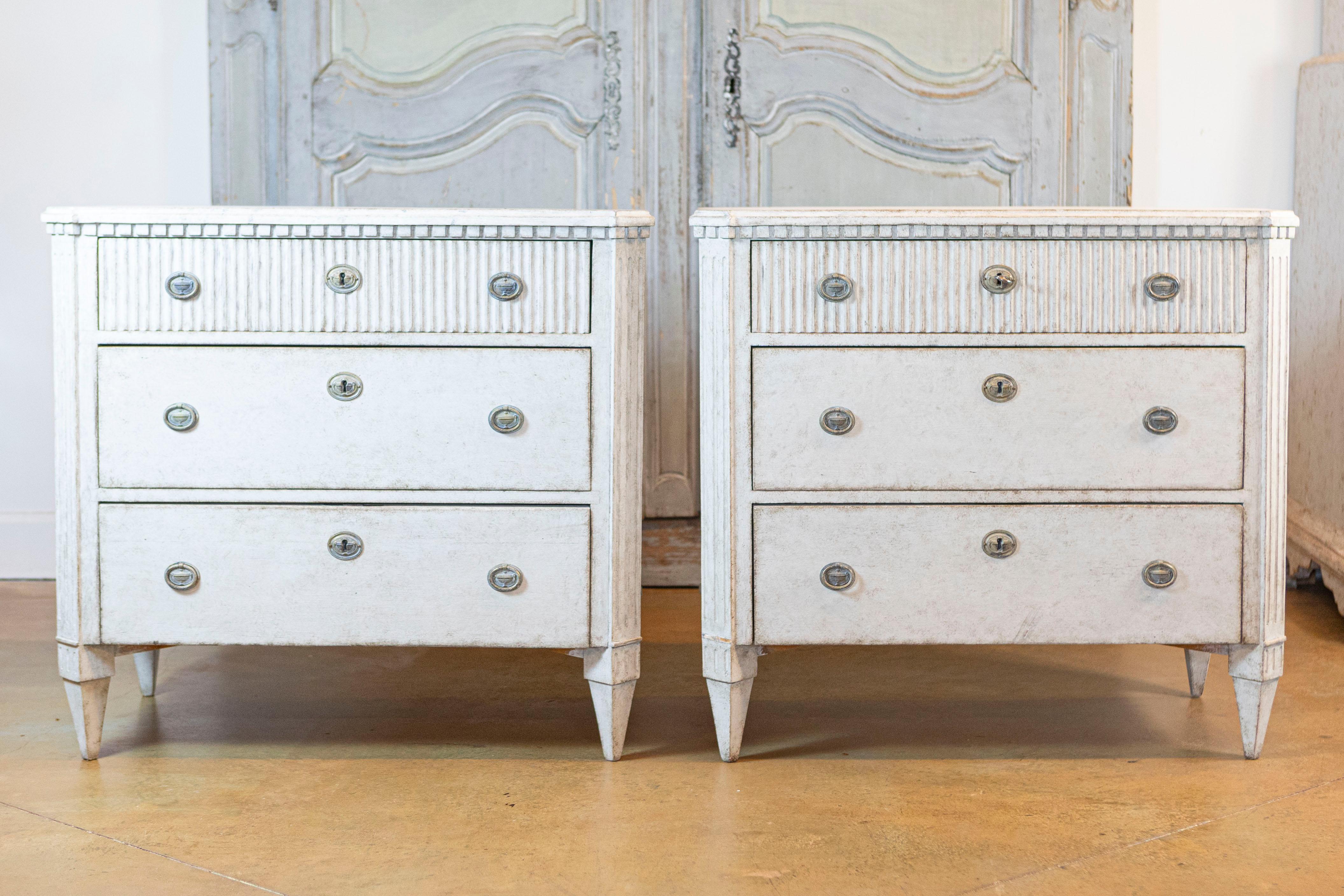A pair of Swedish Gustavian style three-drawer chests from circa 1870 with lightly marbleized tops, gray painted finish and carved reeded motifs. Elevate your interior with this stunning pair of Swedish Gustavian-style three-drawer chests from