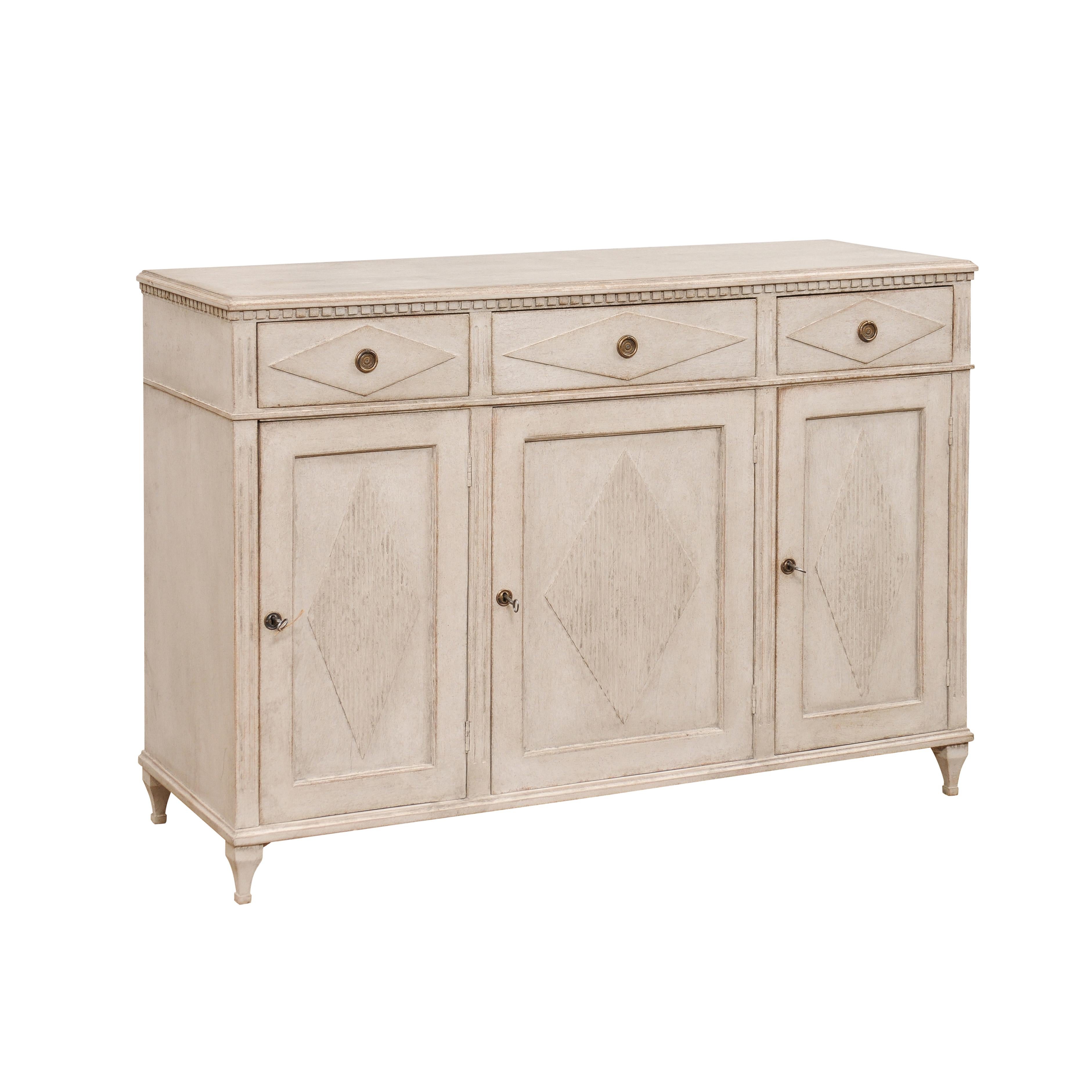 A Swedish Gustavian Style painted wood sideboard circa 1870 with carved dentil molding, three drawers, three doors and carved diamond motifs. Transport yourself to the refined elegance of Swedish Gustavian design with this exquisite painted wood