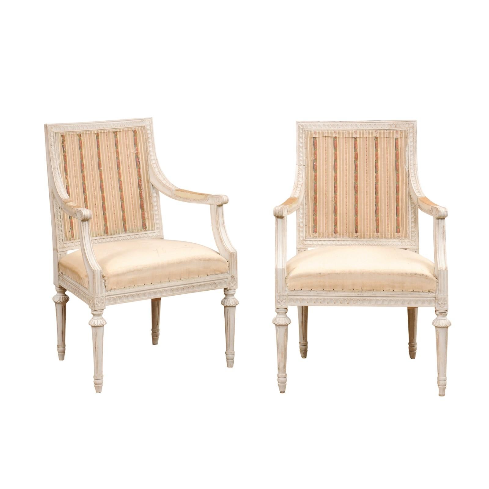 A pair of Swedish Gustavian style painted wood armchairs from the early 20th century, with scrolling arms, carved waterleaf and rosette motifs and fluted legs. Created in Sweden during the early years of the 20th century, each of this pair of