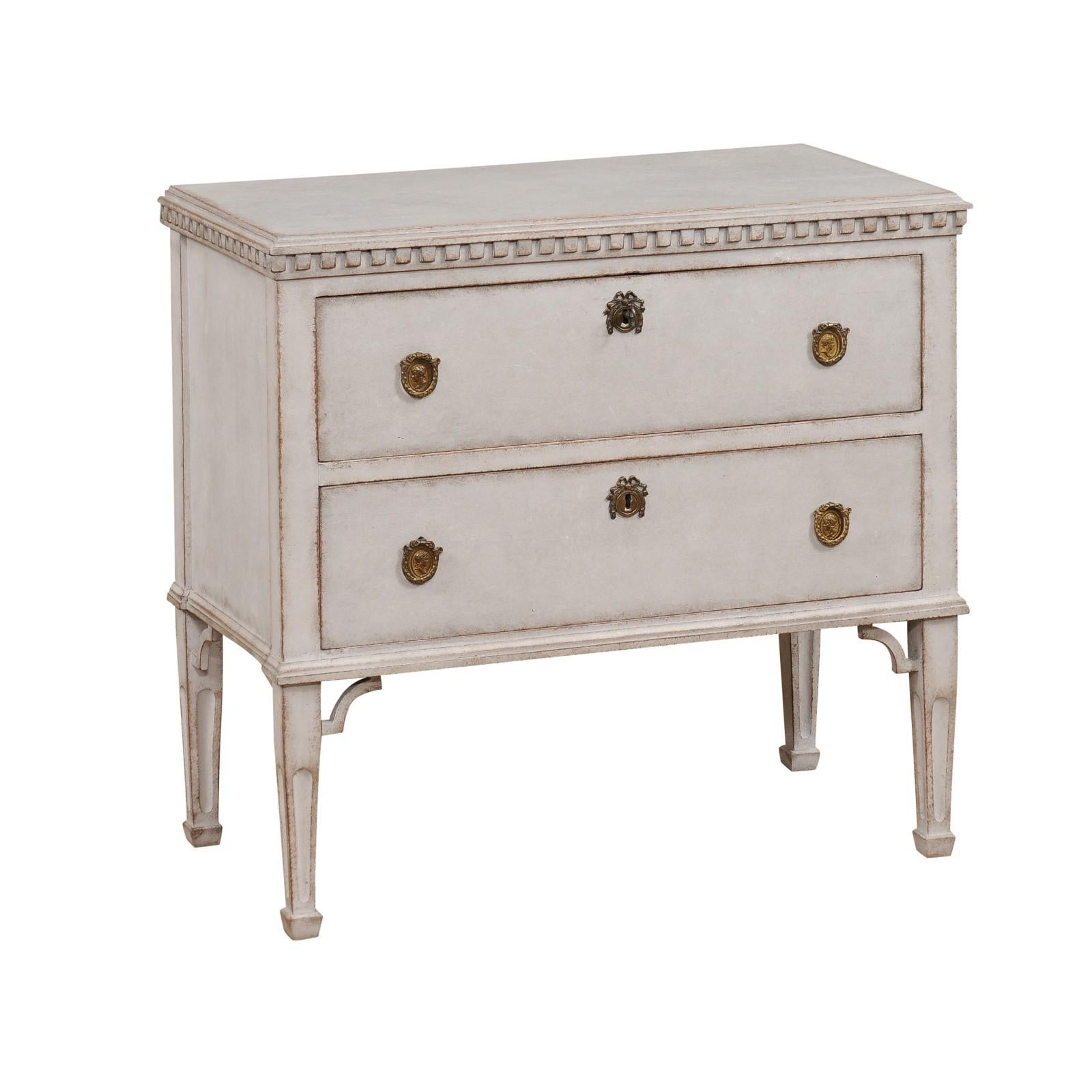 A Swedish Gustavian style chest of drawers from the 19th century with light gray paint, two drawers, carved dentil molding, tapered legs and carved spandrels. This Swedish Gustavian style chest of drawers, hailing from the 19th century, exudes the