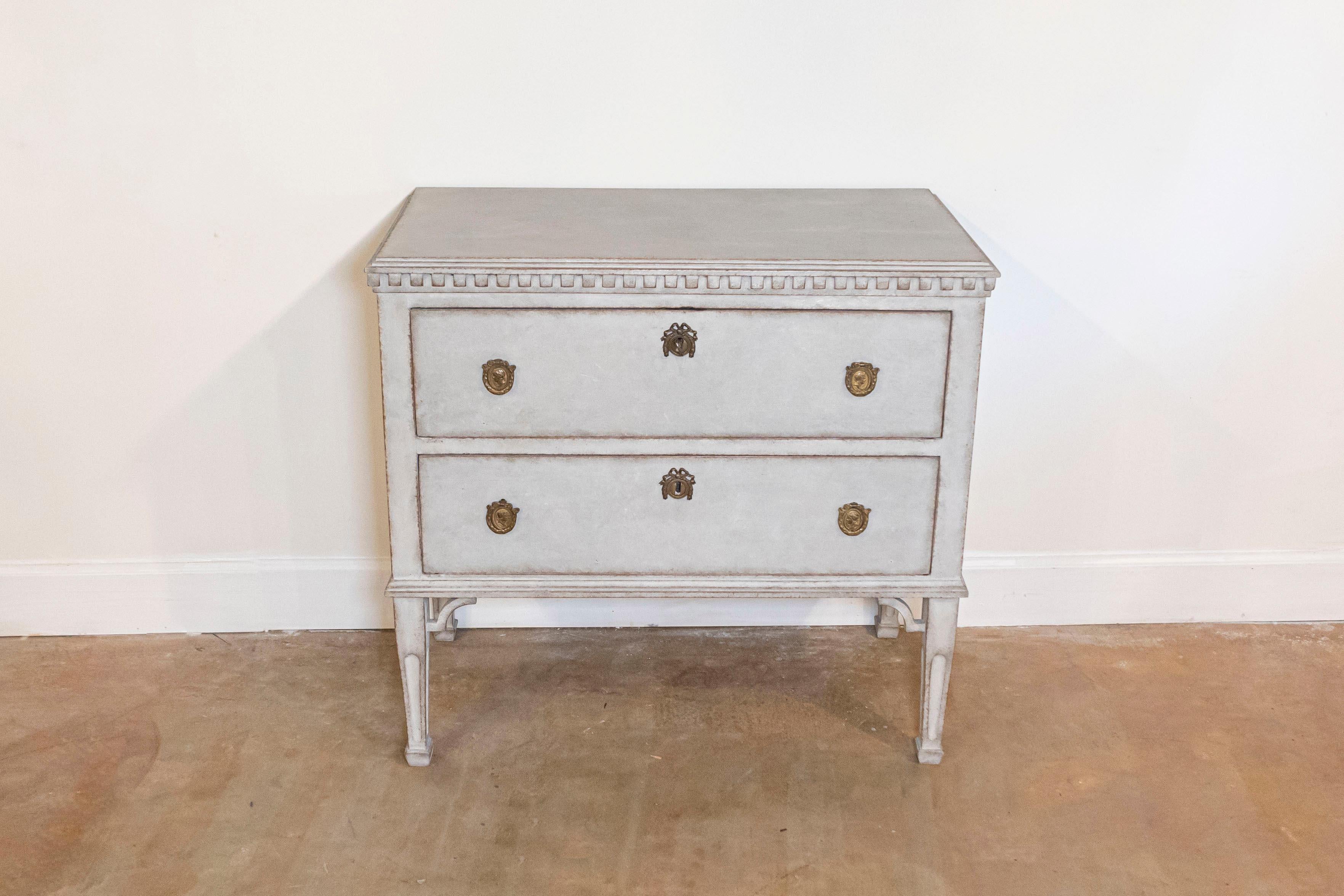 A Swedish Gustavian style chest of drawers from the 19th century with light gray paint, two drawers, carved dentil molding, tapered legs and carved spandrels. This Swedish Gustavian style chest of drawers, hailing from the 19th century, exudes the