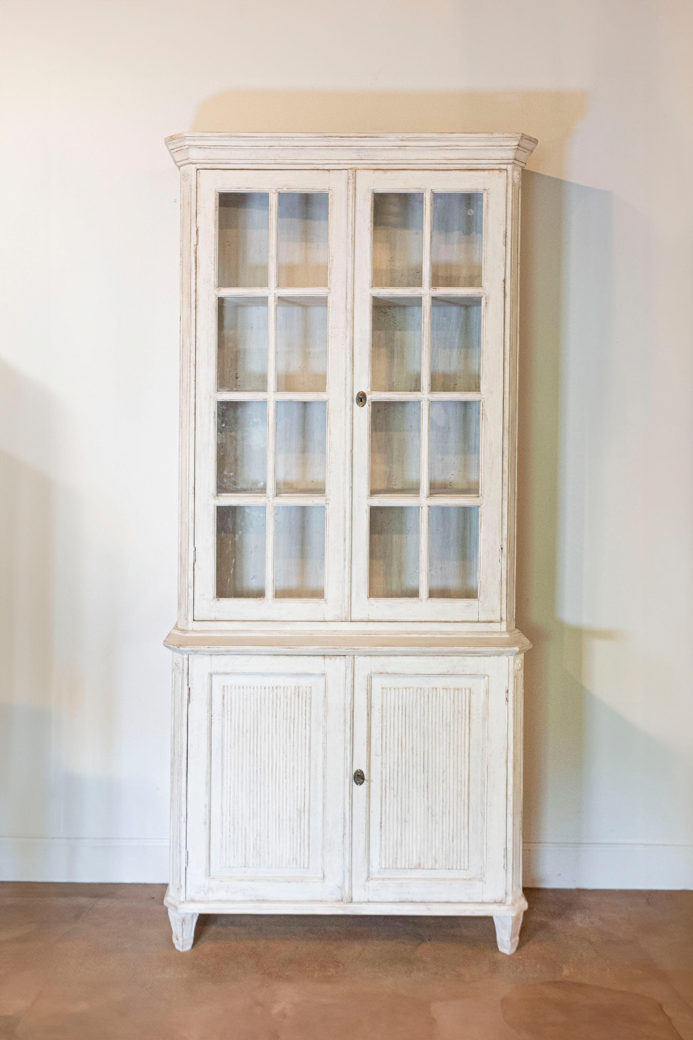 A Swedish Gustavian style two-part vitrine cabinet from the 19th century with light gray / cream painted finish, glass doors, carved reeded panels and tapered feet. Immerse yourself in the timeless elegance of this Swedish Gustavian style two-part