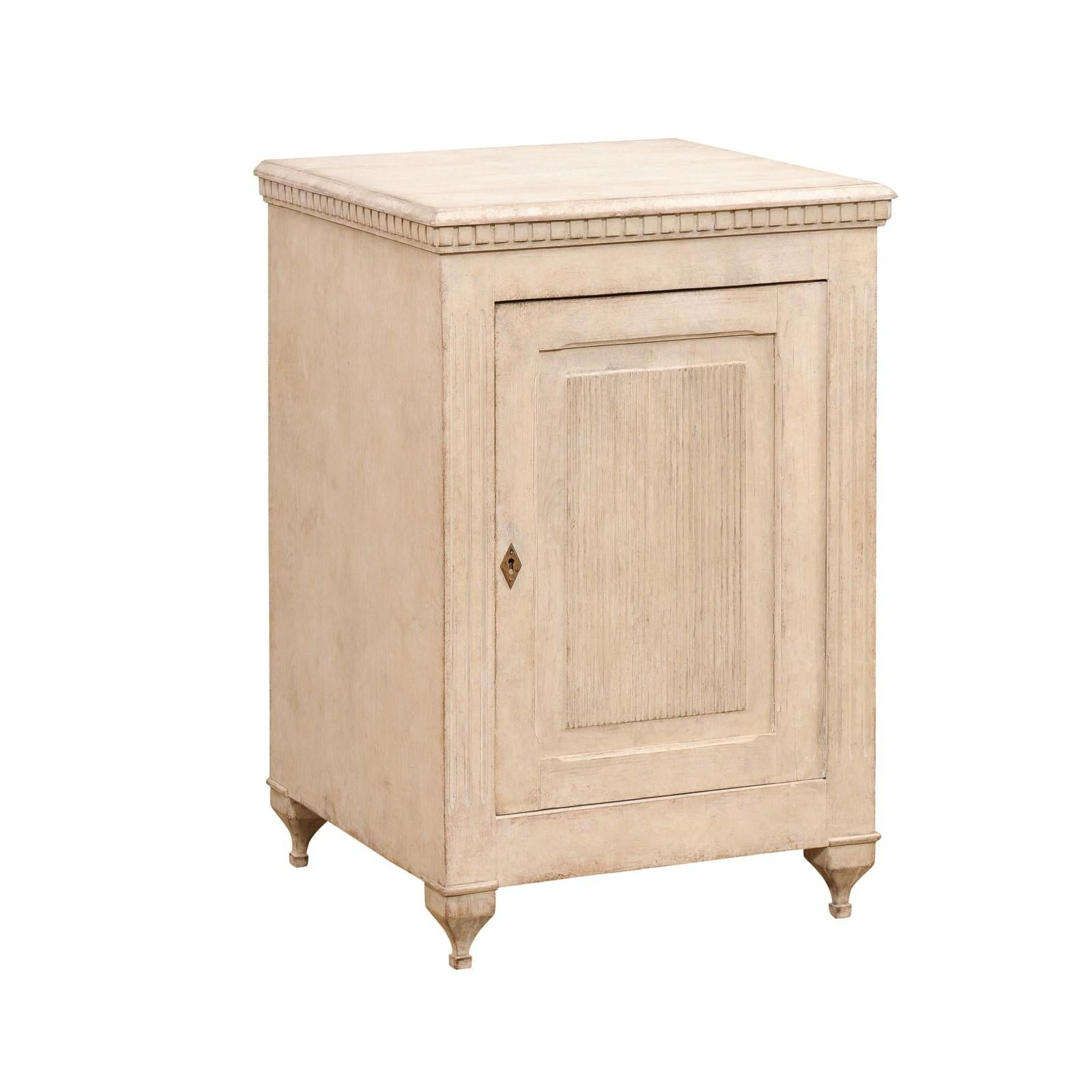 A Swedish Gustavian style painted wood side cabinet from the 19th century, with carved dentil molding, reeded door and petite carved feet. Created in Sweden during the 19th century, this painted side cabinet features the stylistic characteristics of