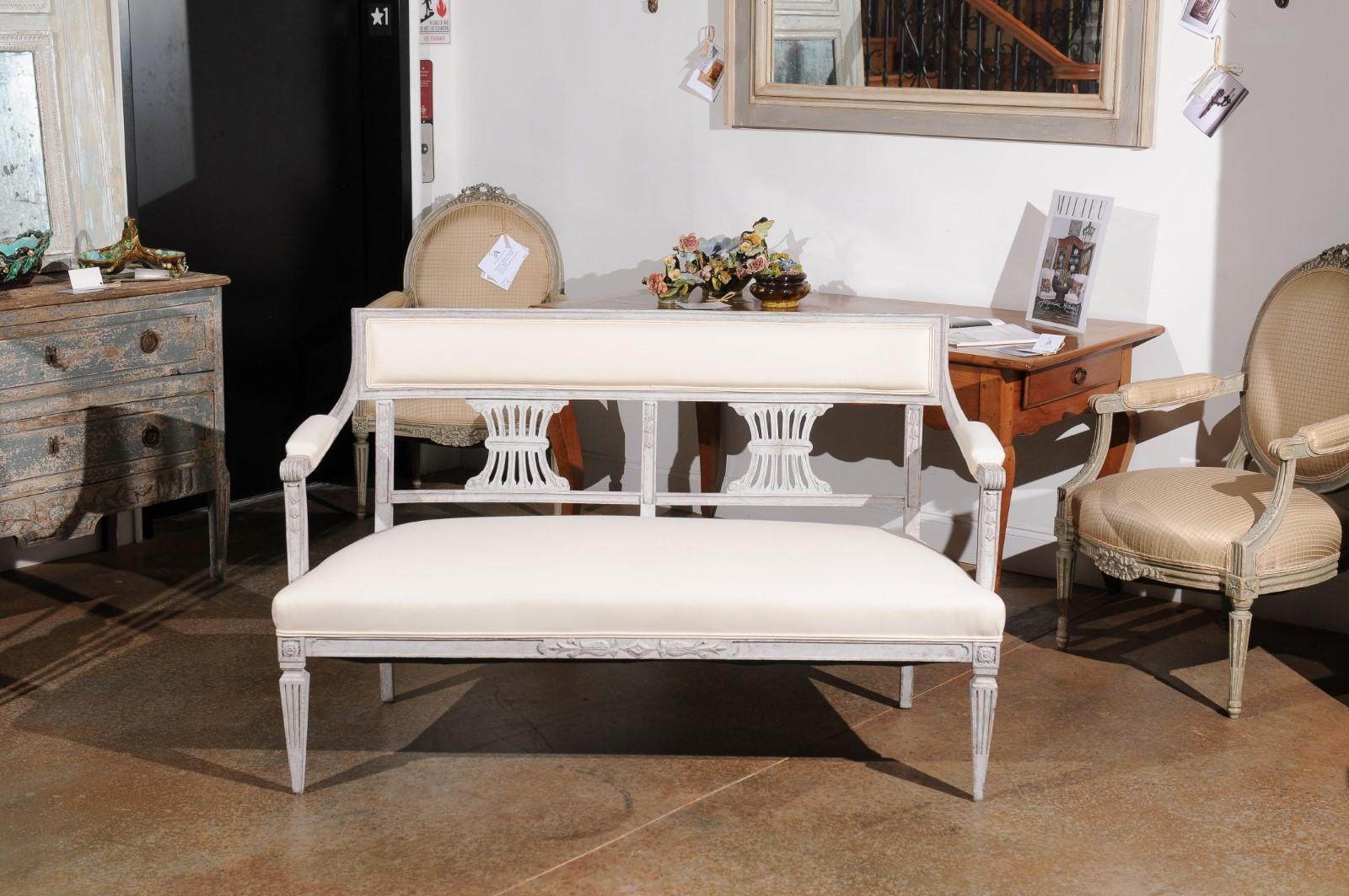 A Swedish Gustavian style painted wood settee bench from the 19th century, with carved back and new upholstery. Created in Sweden during the 19th century, this Gustavian style settee features an open back adorned with carved motifs, connected to two