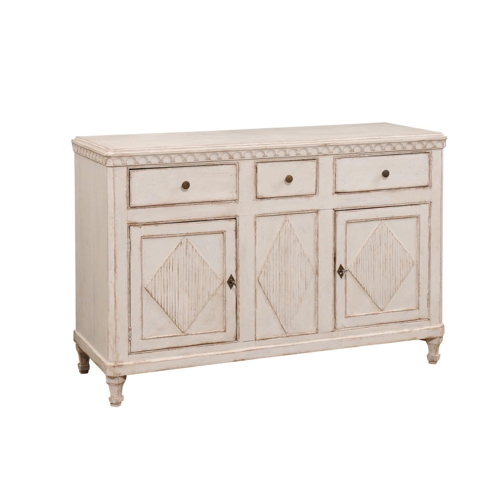 A Swedish Gustavian style painted wood sideboard from the 19th century with three drawers, two doors, carved arched-themed frieze, reeded diamond motifs and tapering feet. This alluring Swedish Gustavian style painted wood sideboard, hailing from
