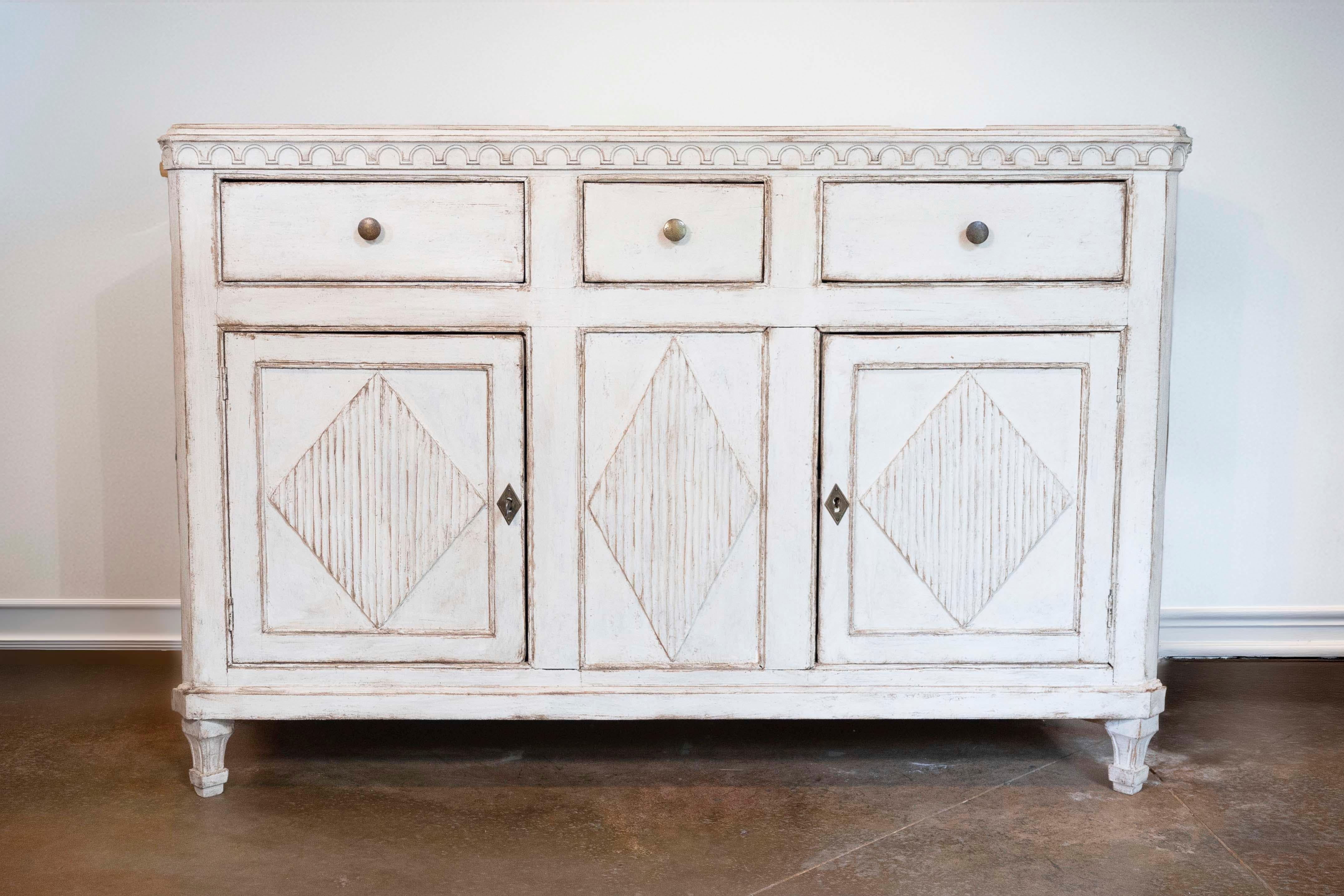 A Swedish Gustavian style painted wood sideboard from the 19th century with three drawers, two doors, carved arched-themed frieze, reeded diamond motifs and tapering feet. This alluring Swedish Gustavian style painted wood sideboard, hailing from