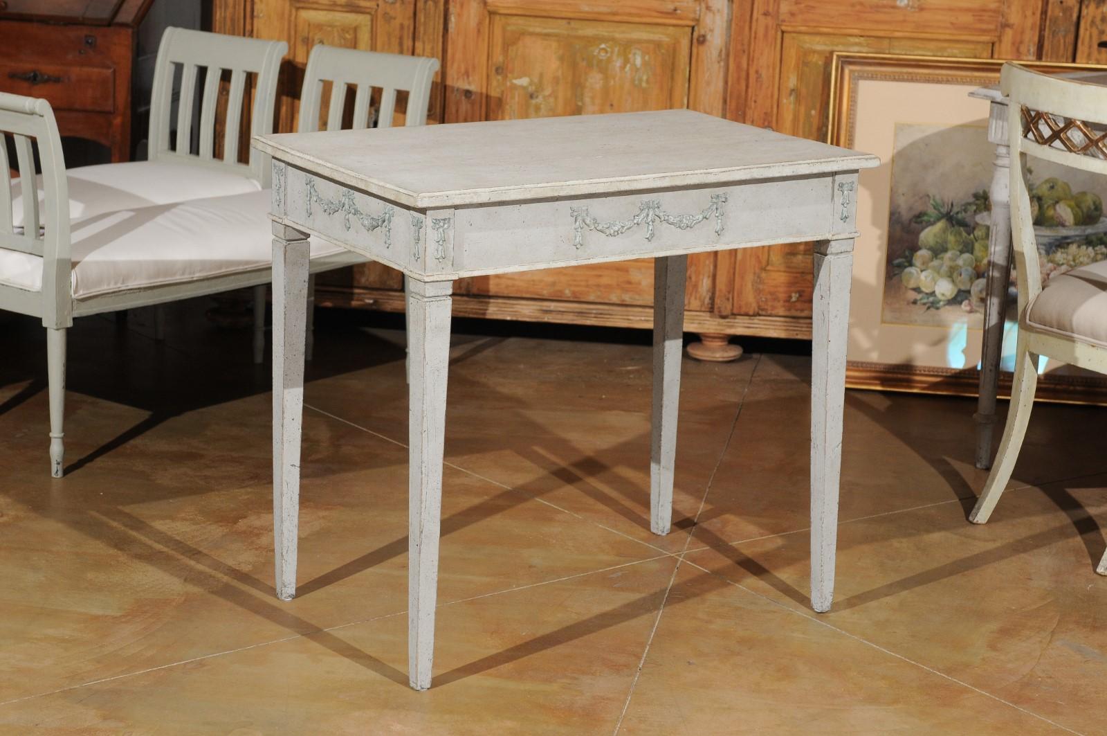 A Swedish Gustavian style painted wood table from the 19th century, with carved swags and tapered legs. Created in Sweden during the 19th century, this painted table features a rectangular top sitting above an elegant apron adorned with carved