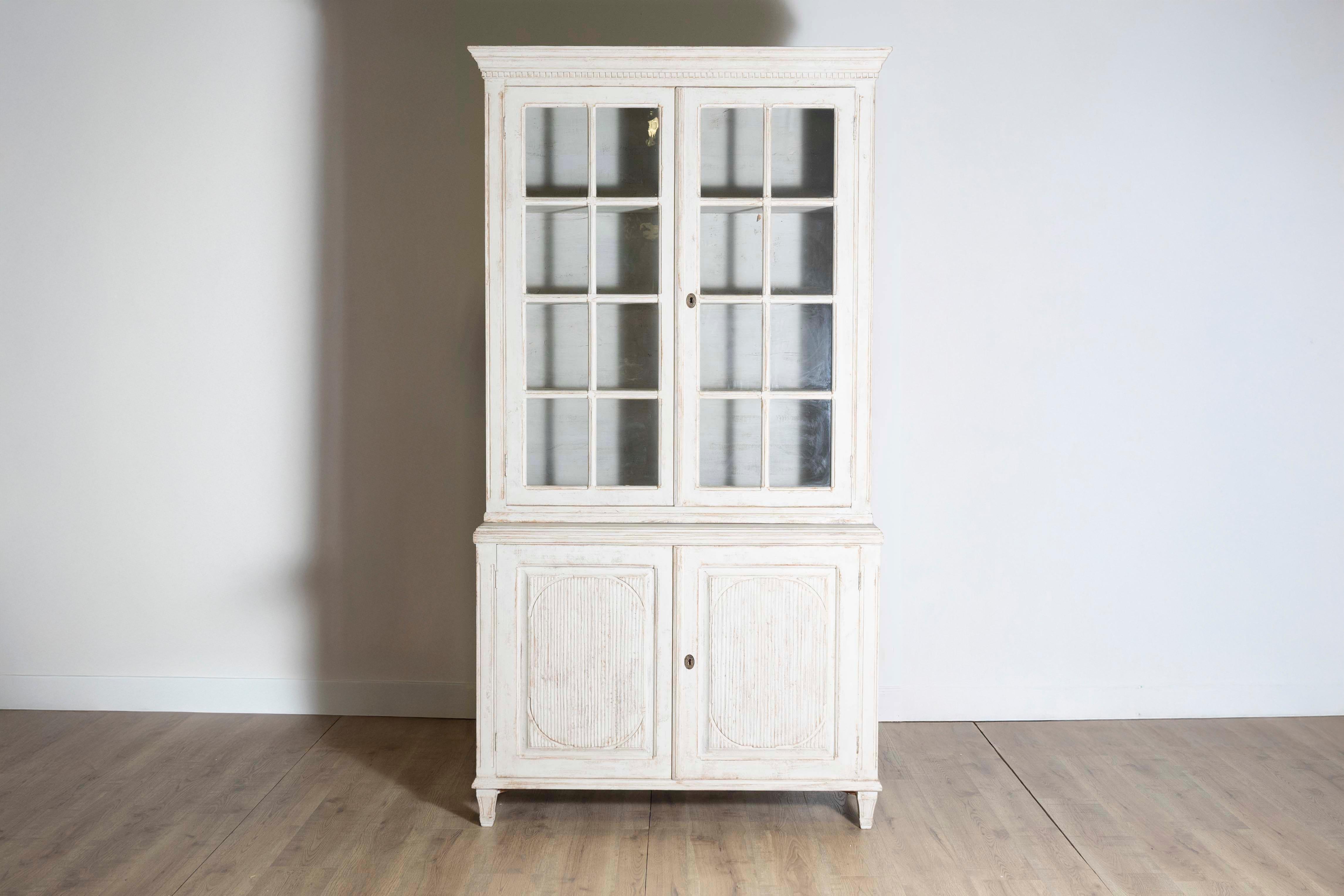 A Swedish Gustavian style painted wood vitrine cabinet from the 19th century with glass doors, carved reeded panels and tapered feet. This Swedish Gustavian style painted wood vitrine cabinet from the 19th century, embodies the sophisticated
