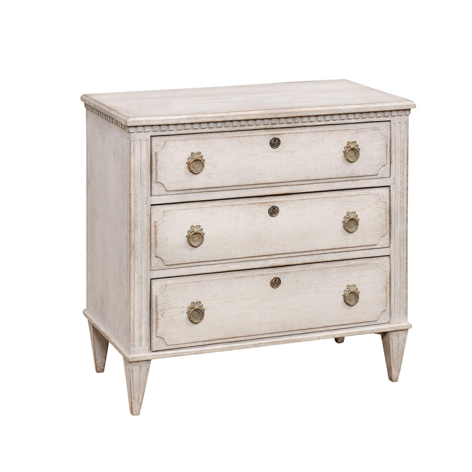 A Swedish Gustavian style painted wood chest from the 19th century, with three drawers, carved dentil molding, fluted side supports and tapered feet. Created in Sweden during the 19th century, this chest of drawers showcases the stylistic