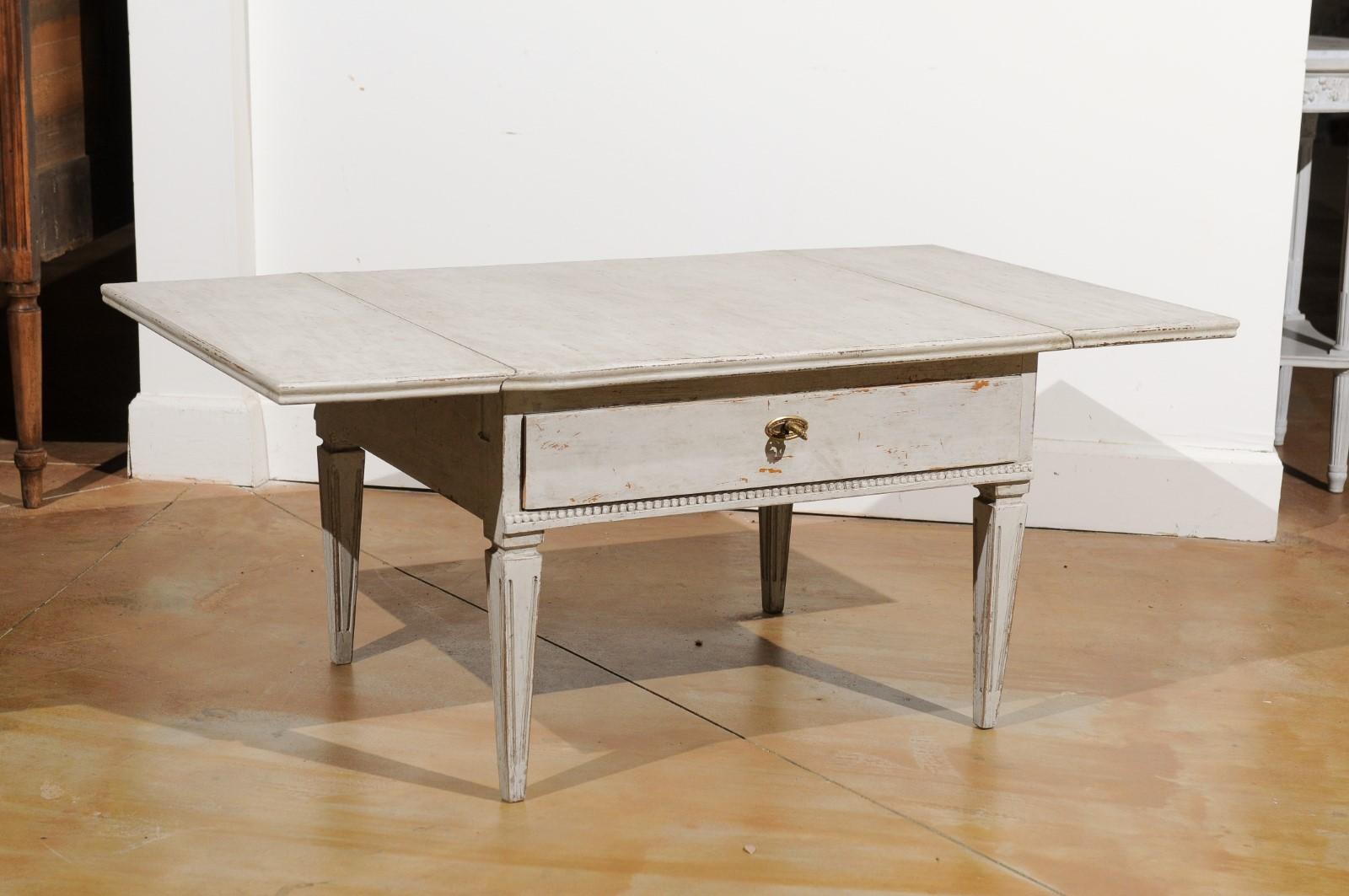 A Swedish Gustavian style painted coffee table from the 20th century, with drop leaves, single drawer and tapered legs. Created in Sweden during the 20th century, this Gustavian style coffee table features a rectangular top flanked with drop leaves,
