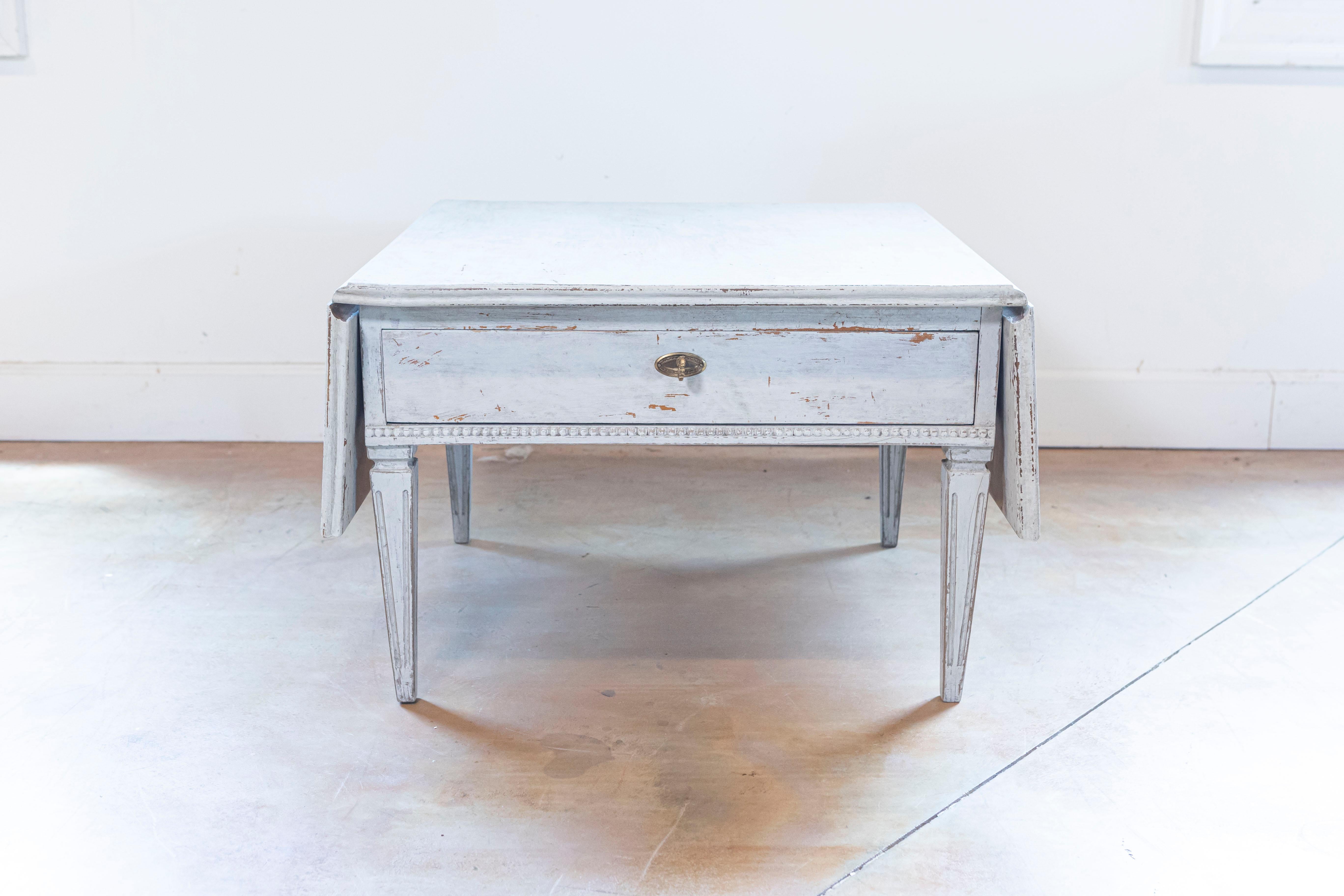 A Swedish Gustavian style painted coffee table from the 20th century, with drop leaves, single drawer and tapered legs. Created in Sweden during the 20th century, this Gustavian style coffee table features a rectangular top flanked with drop leaves,
