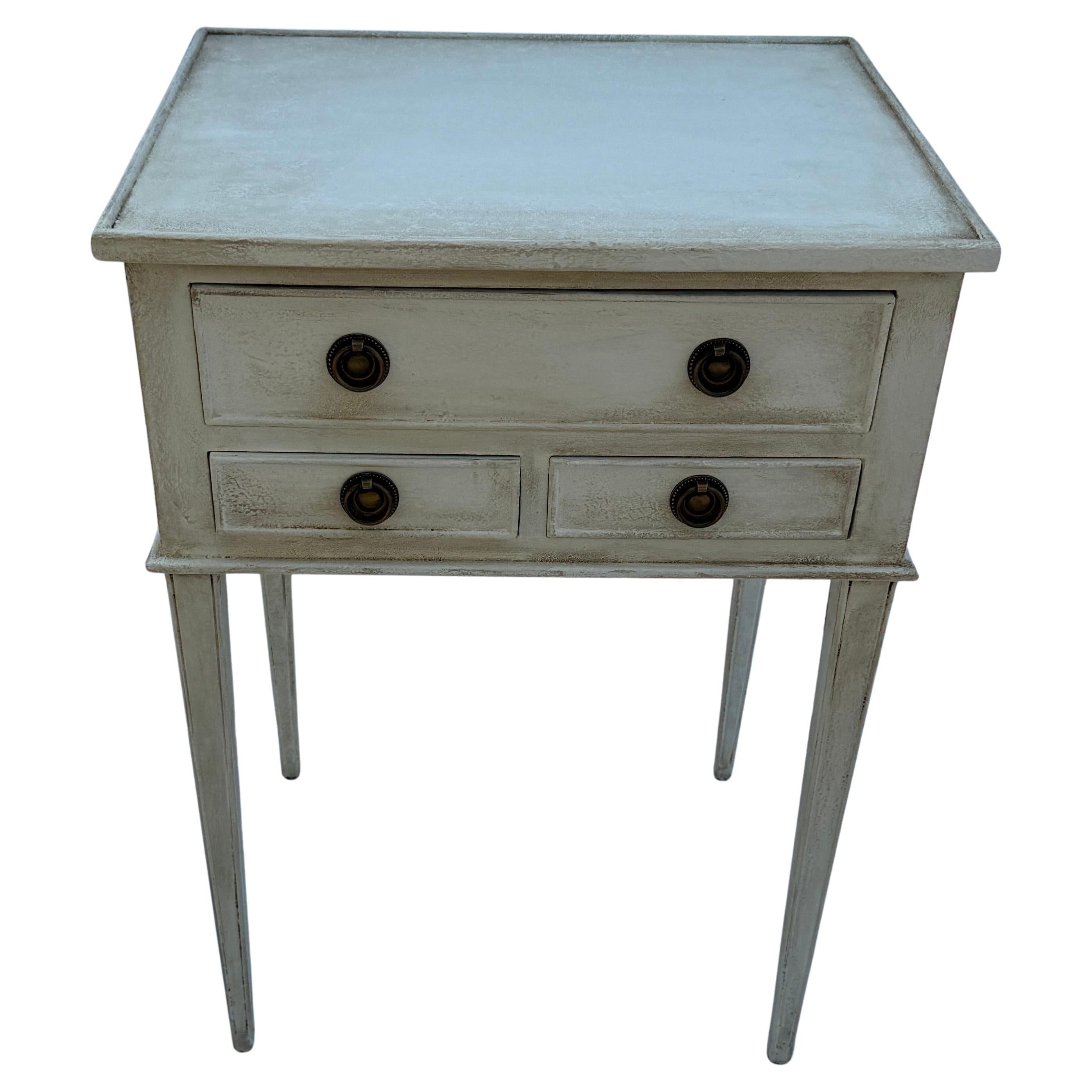 This Swedish style three drawer hand painted neutral toned bedside table has been constructed from solid wood. Classic details on this table including a hand-applied finish and feature bronzed ring hardware. This piece has been based on an 18th