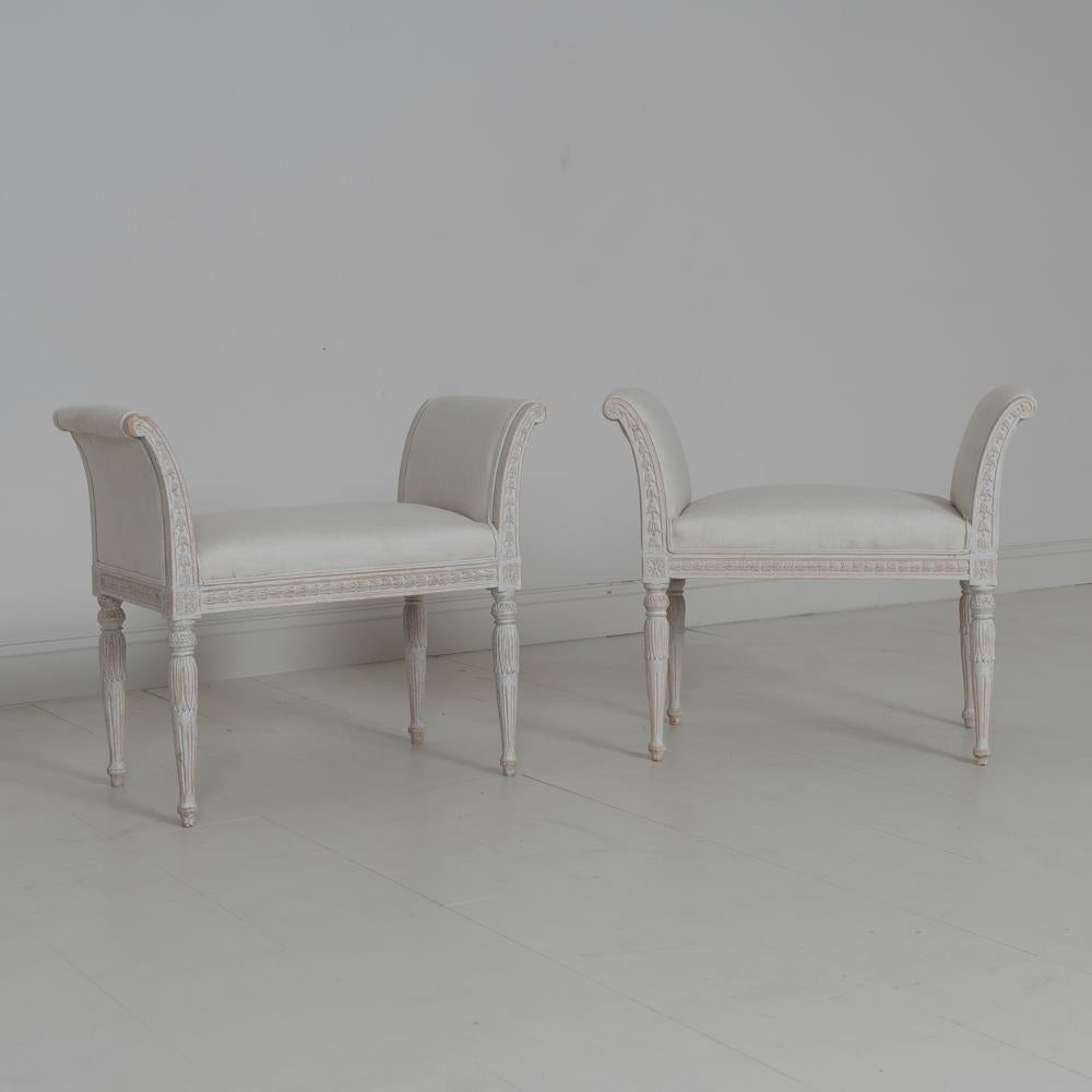 A beautiful pair of Swedish Gustavian style stools with upholstered arms, newly upholstered in linen. There are carved bell flowers around the seat frame and arms with carved rosettes on the corner posts. The round and tapered legs are embellished