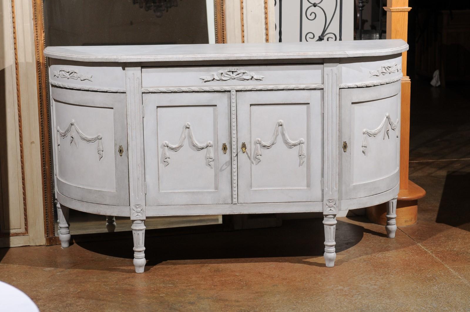 A Swedish Gustavian style painted wood demilune sideboard from the 20th century with carved swags and ribbons. Born in Sweden during the 20th century, this exquisite sideboard features an elongated semi-circular top sitting above a lovely breakfront