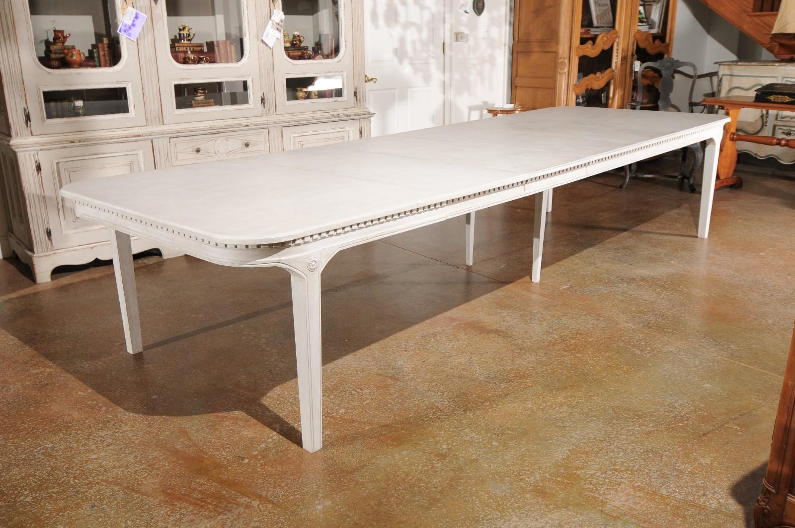 A Swedish Gustavian style painted wood extension dining table from the 20th century, with four leaves, dentil molding and tapered legs. Born in Sweden during the 20th century, this exquisite dining table features a rectangular top with rounded