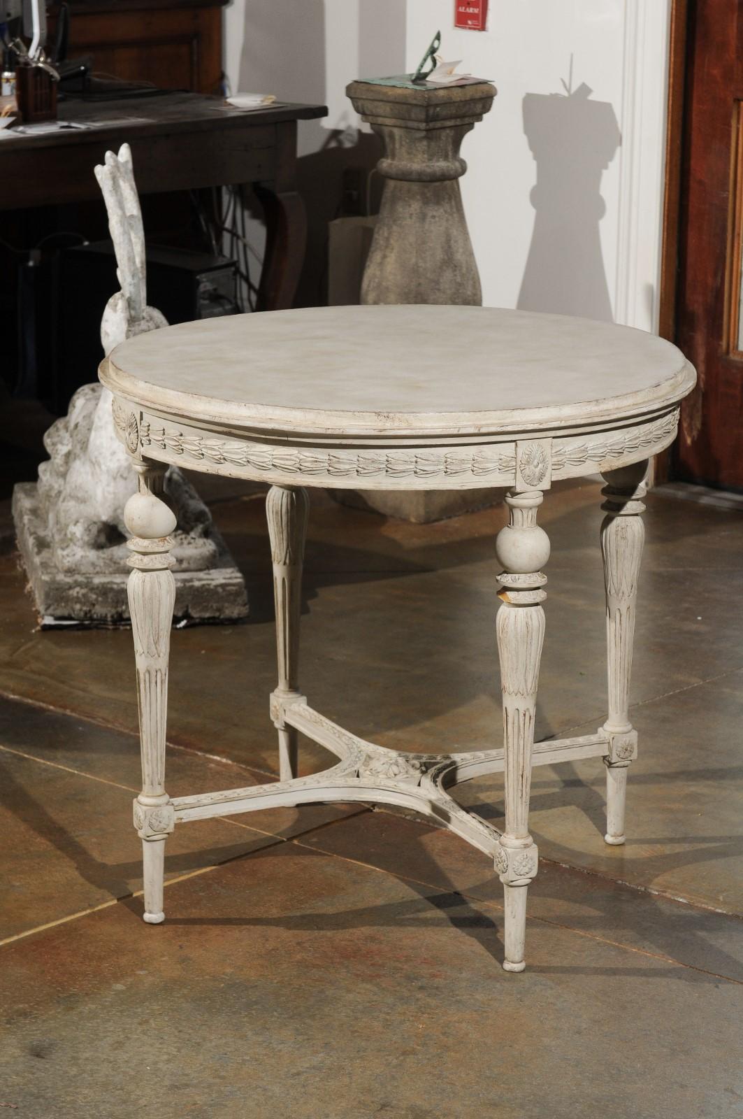 A Swedish Gustavian style painted wood round table from the 19th century, with carved apron, fluted legs and X-form cross stretcher. Born in Sweden during the 19th century, this elegant table charms us with its Gustavian inspired lines. Featuring a