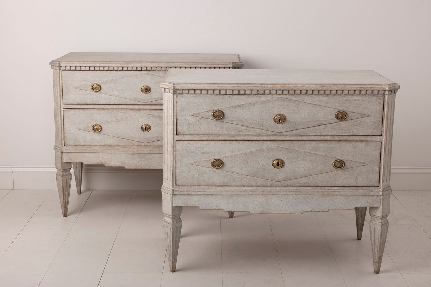 A pair of Swedish commodes in the Gustavian style. These chests feature a raised lozenge design on the drawer fronts with dentil molding around the tops of the commodes. Two large drawers, brass hardware, canted and fluted corner posts, and tapered