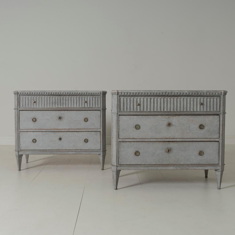 A beautiful pair of Swedish commodes in the Gustavian style with hand painted marbleized tops. There is dentil molding around the top with a fluted top drawer. Chalky gray paint. Three drawers, canted and fluted corner posts, and tapered and fluted